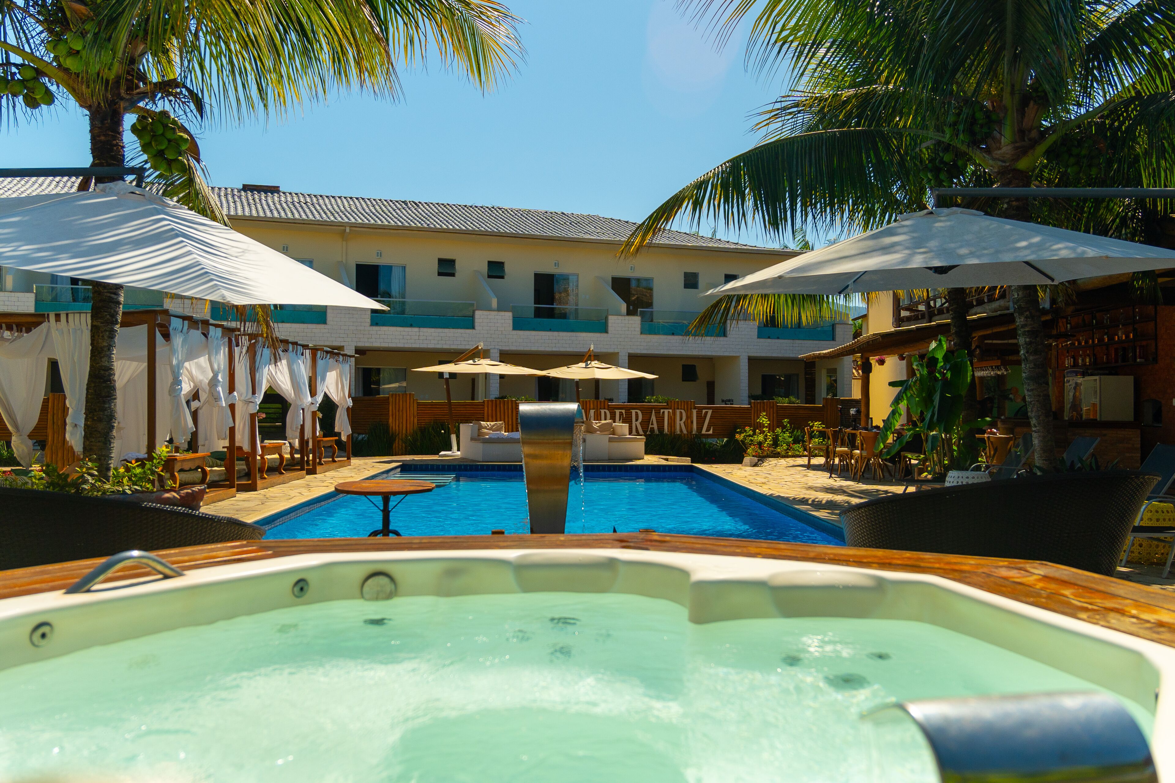 Zona de piscina con sillones de descanso y sombrillas, un jacuzzi y un edificio con balcones.