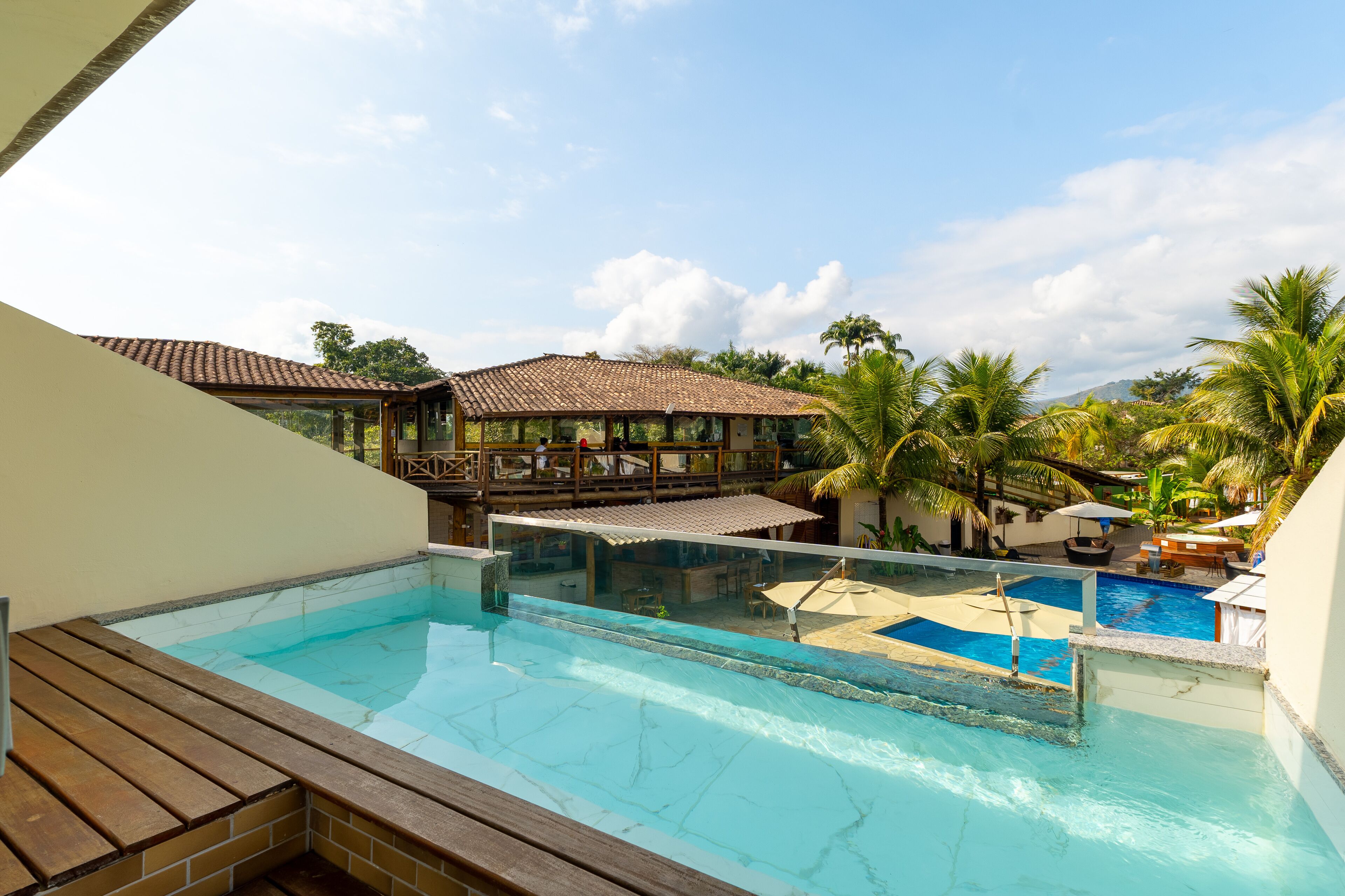 Ein Pool mit Infinity-Kante auf dem Dach, mit Holzterrasse, überdachtem Sitzbereich und Blick auf ein Resorthaus mit Ziegeldach.