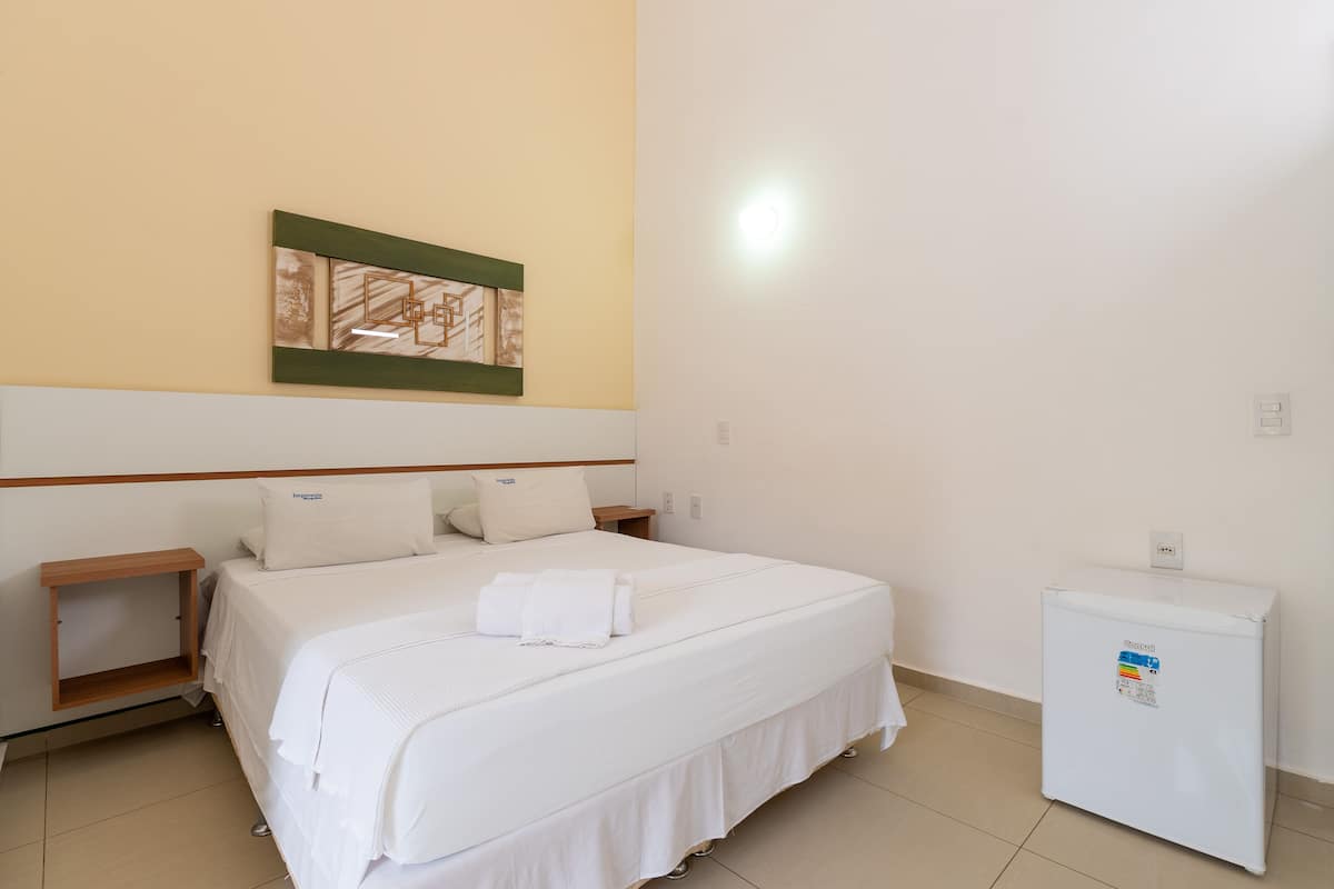 Una habitación de hotel con cama, mesita de noche, un cuadro colgado en la pared y un refrigerador.