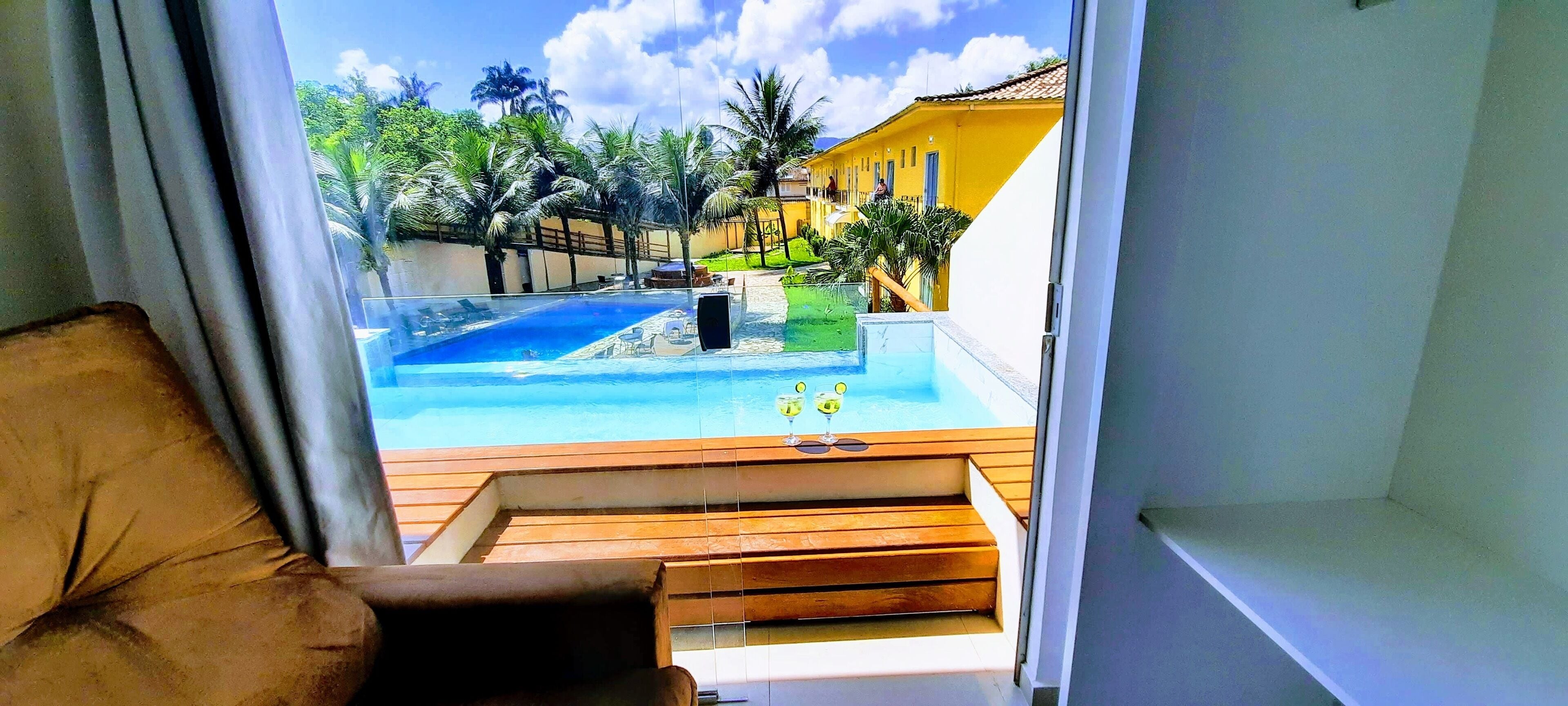 Vista desde una habitación con terraza de madera que da a una piscina, rodeada de vegetación y un edificio amarillo.