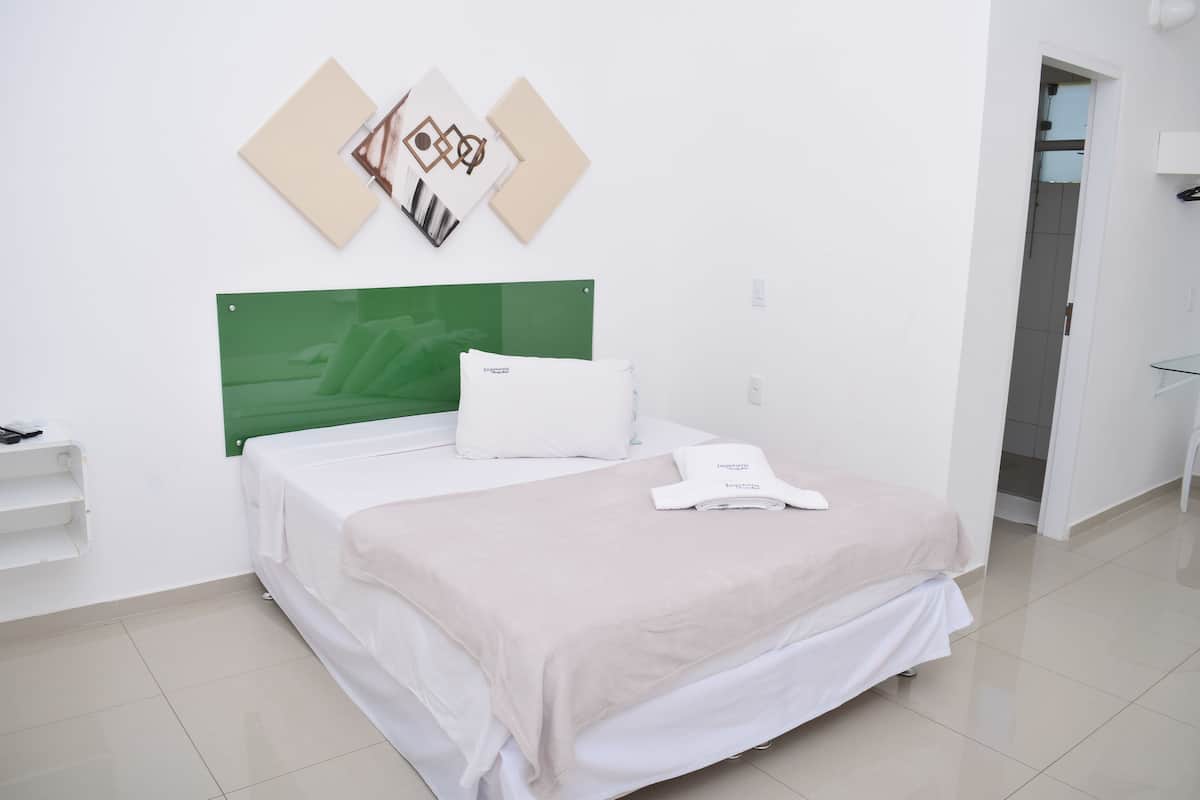 Una cama bien hecha con un cabecero verde, un almohadón blanco y una manta blanca. Hay dos cuadros en la pared sobre la cama.