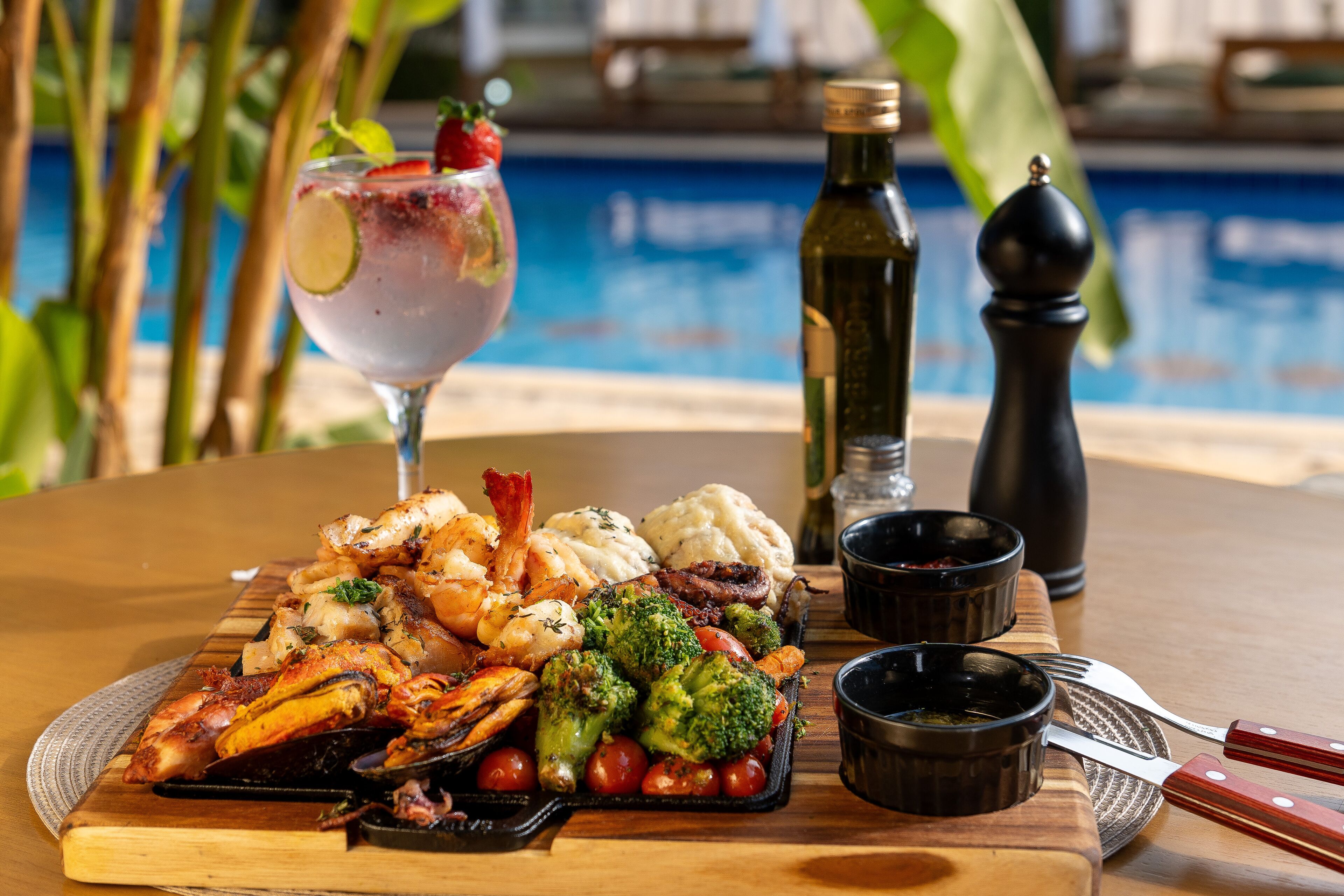 A dining table with a seafood platter, a cocktail, and condiments by a poolside.