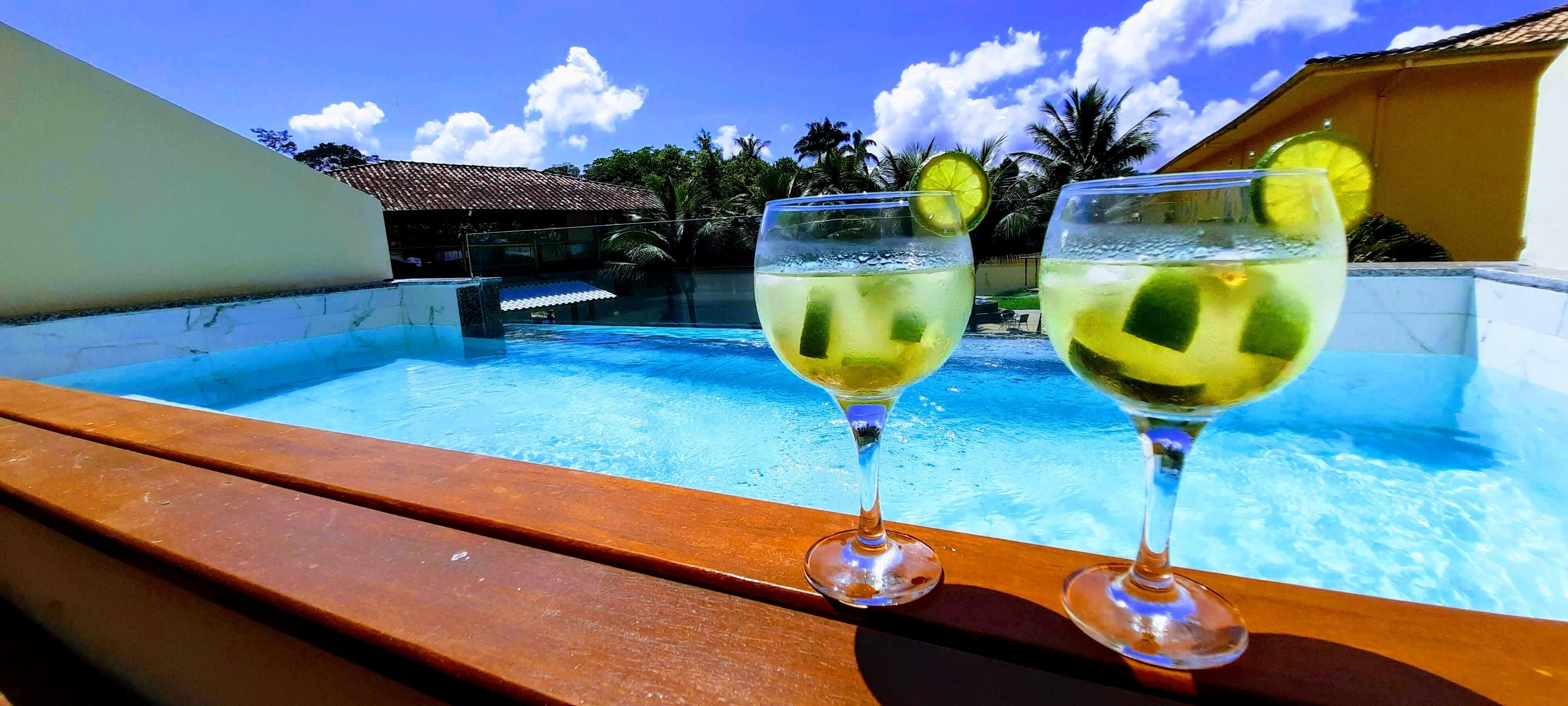 Dos vasos de una bebida clara con rodajas de limón en una terraza de madera con vista a una piscina.