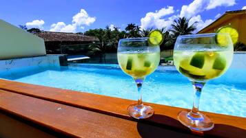 Dos vasos de una bebida clara con rodajas de limón en una terraza de madera con vista a una piscina.