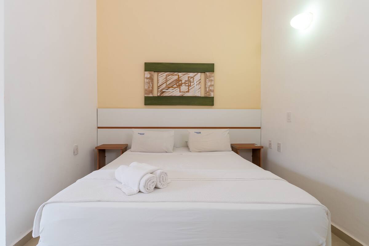 Una cama bien hecha con sábanas blancas y una toalla enrollada, un cabecero de madera y una obra de arte en la pared encima.