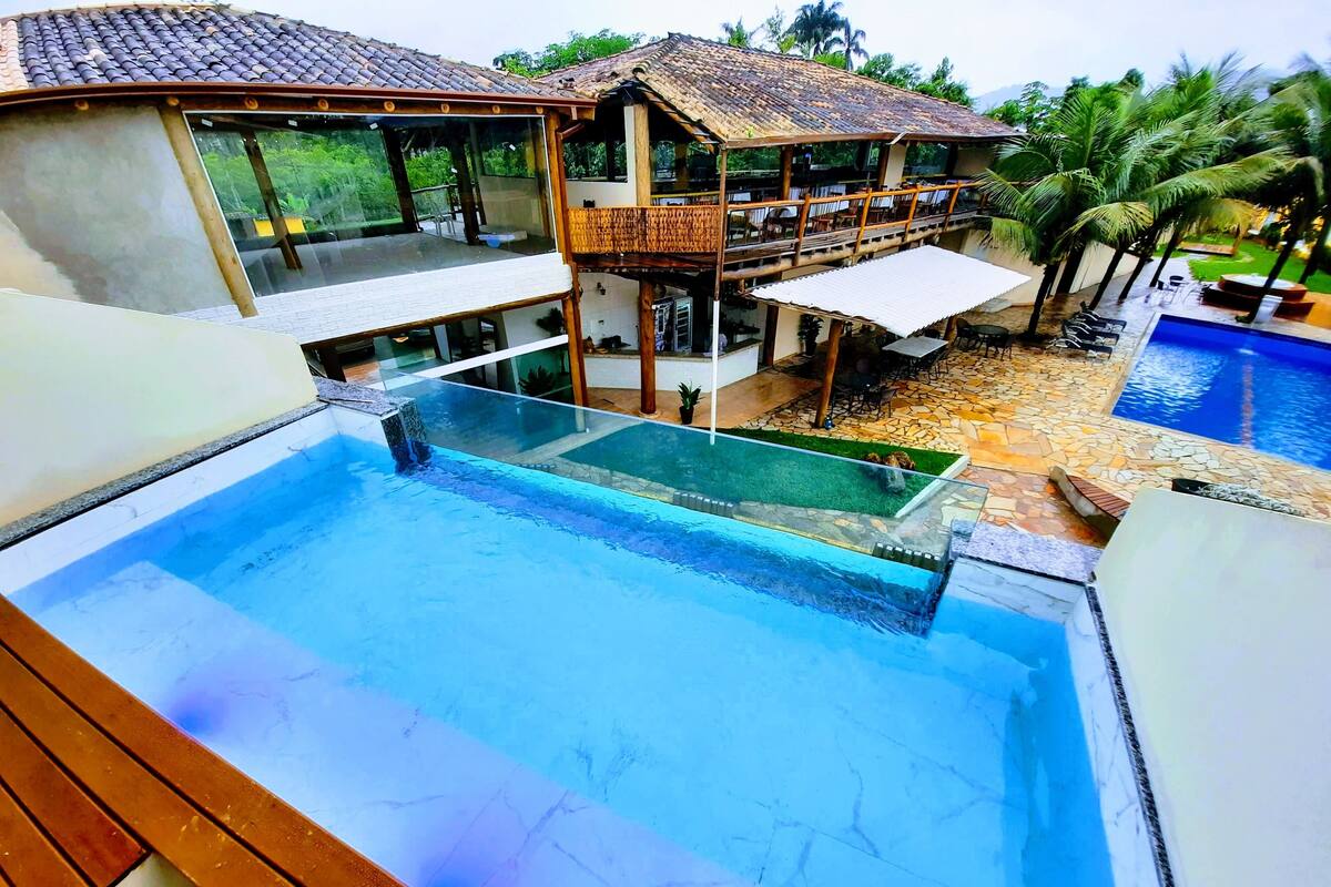 A modern villa with a glass-enclosed infinity pool, a second-story balcony, and a surrounding garden.