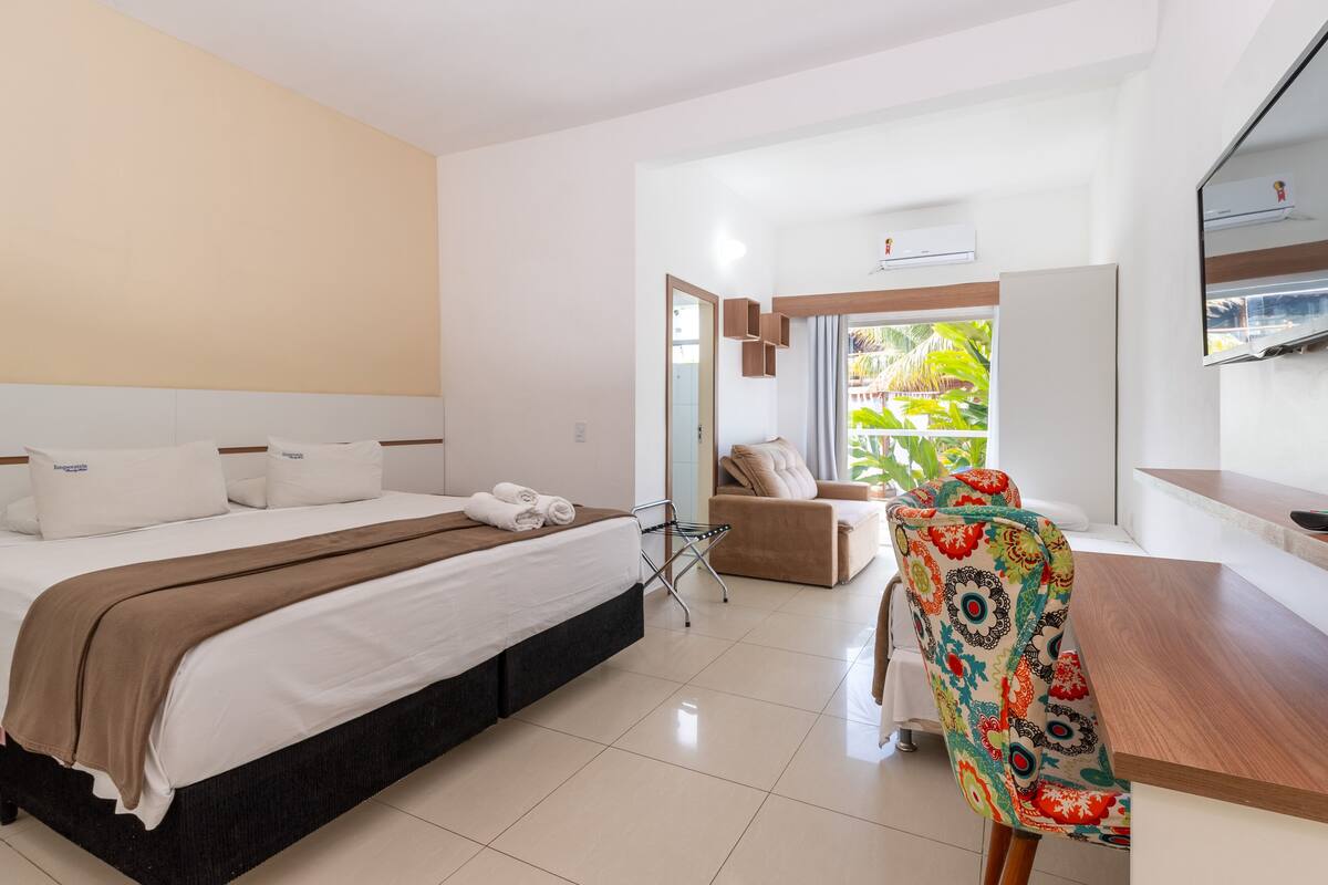 A hotel room with a bed, a chair, a table, and a window with a view of greenery.