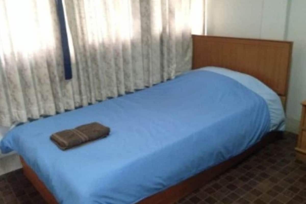 A single bed with a blue bedspread and a brown towel, a wooden headboard, and a window with curtains.