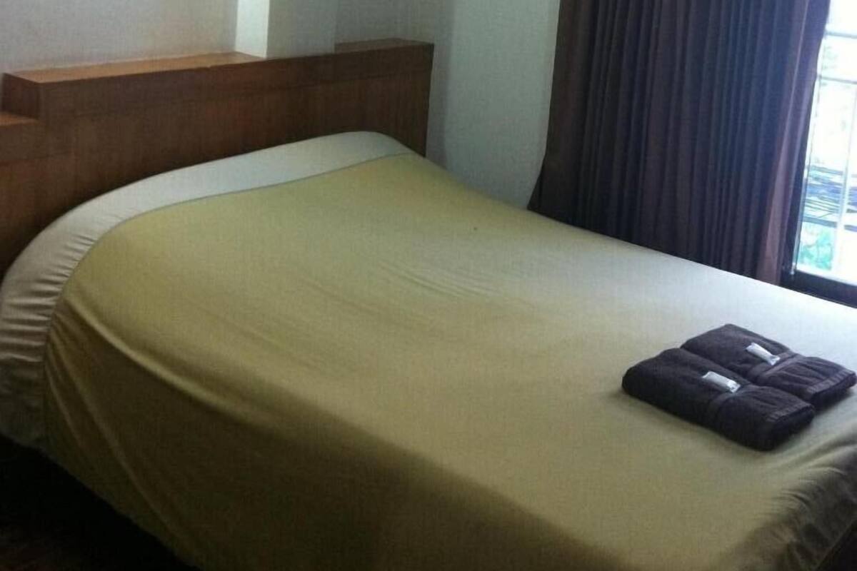A single bed with a wooden headboard, a plain wall, and a window with curtains.