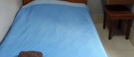 Un lit simple avec un matelas bleu et une serviette marron pliée au pied du lit. Une table de chevet en bois avec une lampe et une banane se trouve à côté du lit.