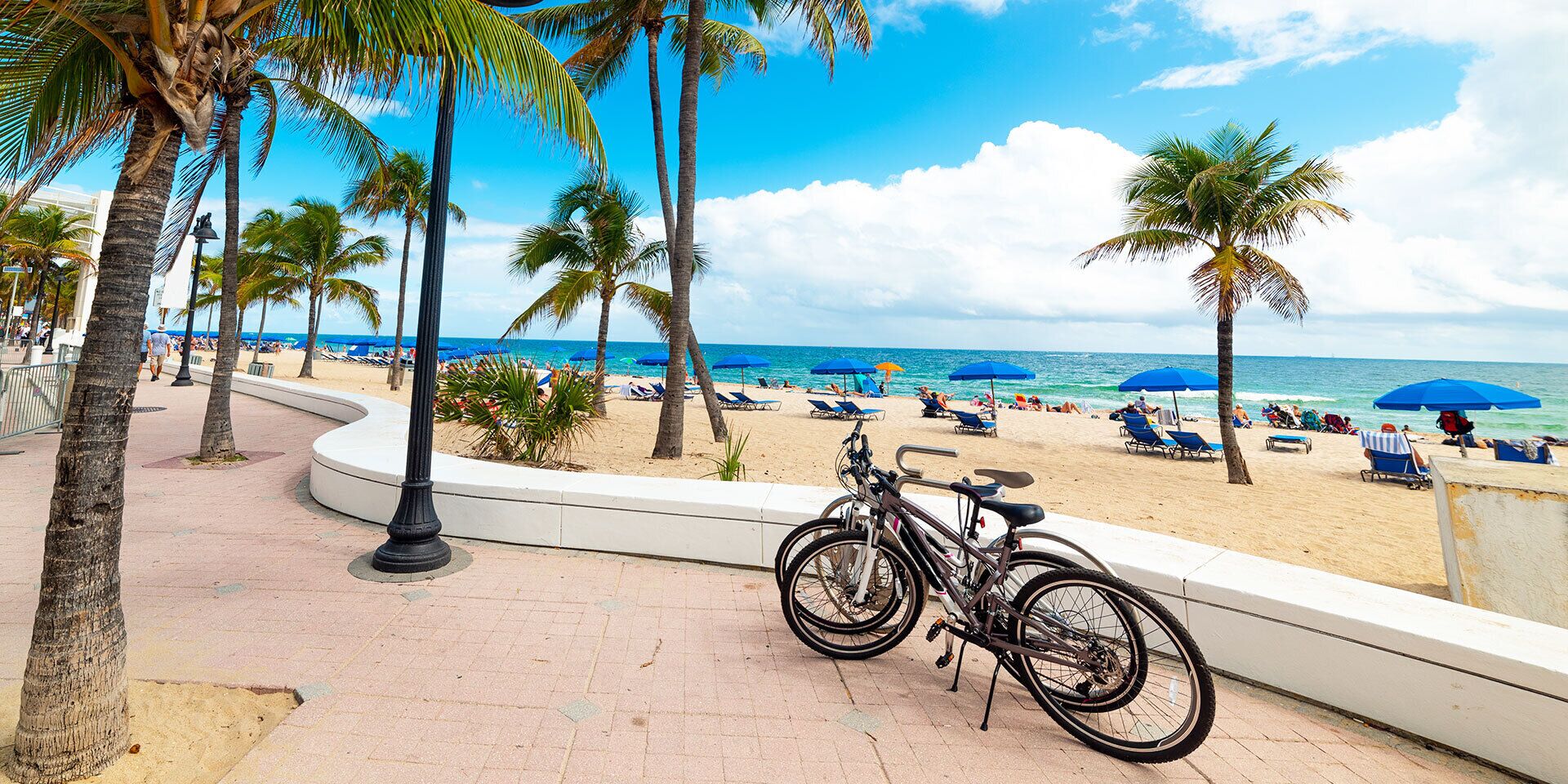 A beachside promenade with bicycles parked, palm trees, and beach umbrellas.