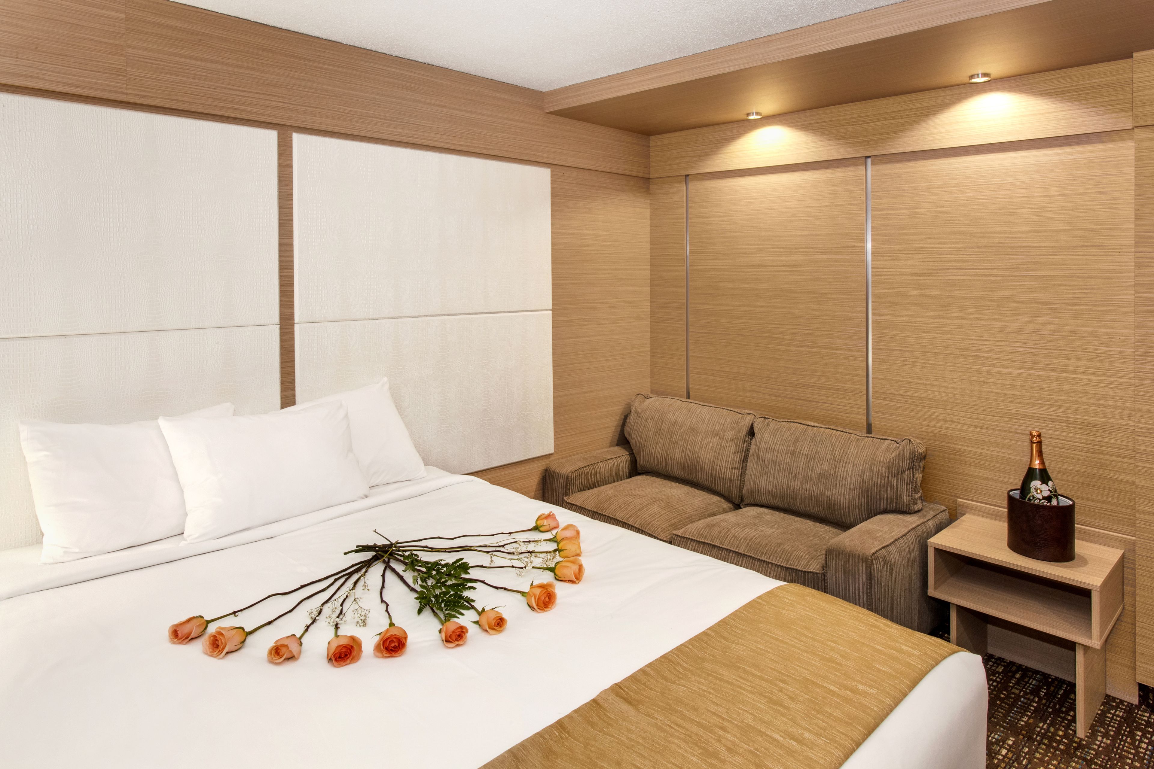 A hotel room with a large bed, a sofa, a nightstand with a bottle of champagne, and a bouquet of flowers on the bed.