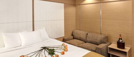 A hotel room with a large bed, a sofa, a nightstand with a bottle of champagne, and a bouquet of flowers on the bed.
