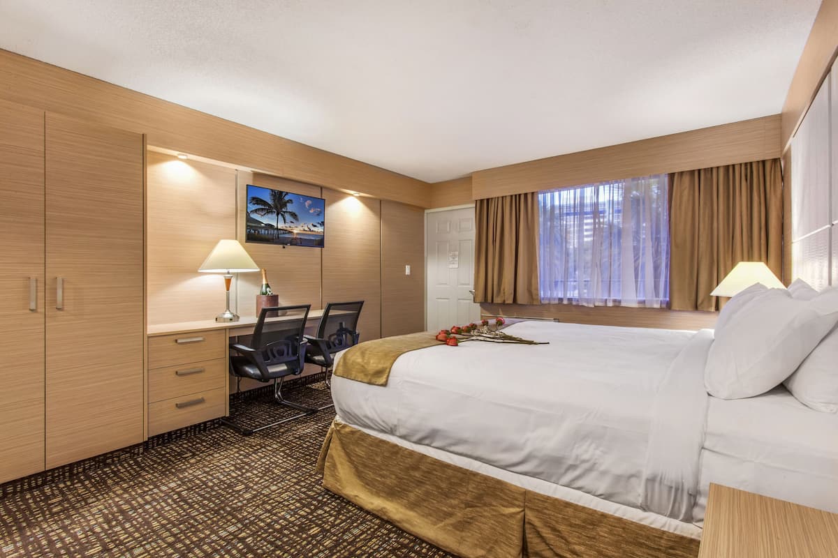 Ein Hotelzimmer mit einem großen Bett, einem Schreibtisch mit Stuhl, einer Schreibtischlampe, einem Fenster mit Vorhängen und einem am Wand montierten Fernseher.