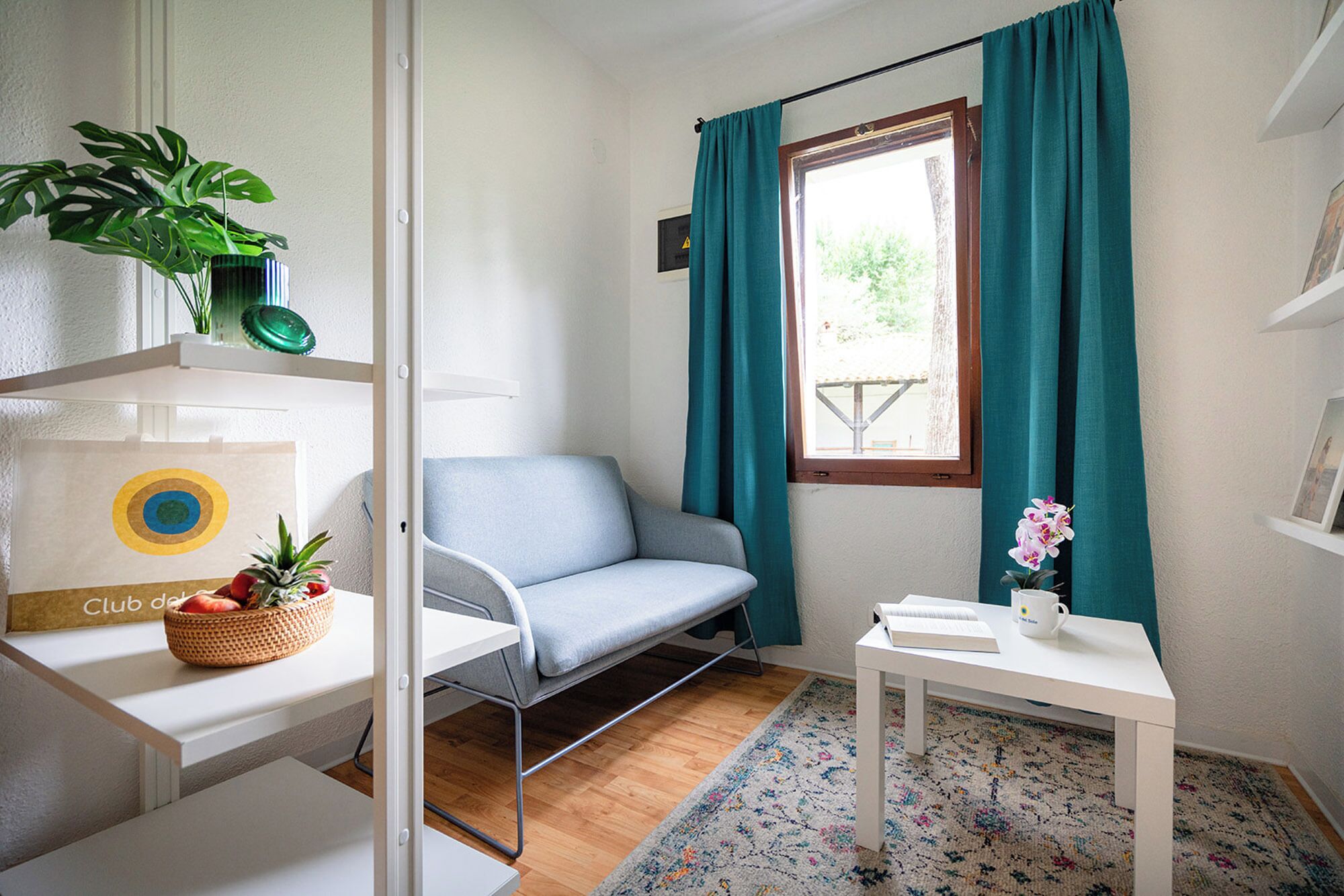 A room with a bed, a chair, a small table, a window with curtains, a plant, and a rug.