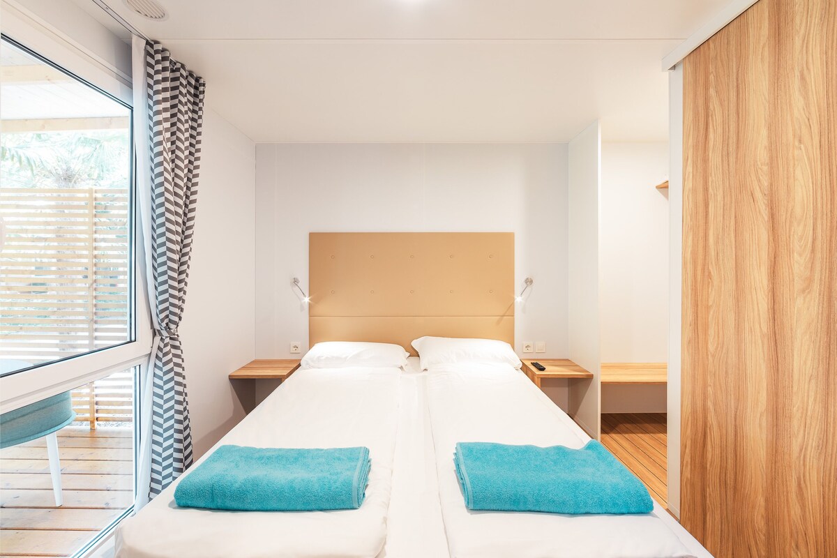 Une chambre double avec deux lits, une armoire en bois, une table de chevet avec une lampe et une fenêtre avec des stores.