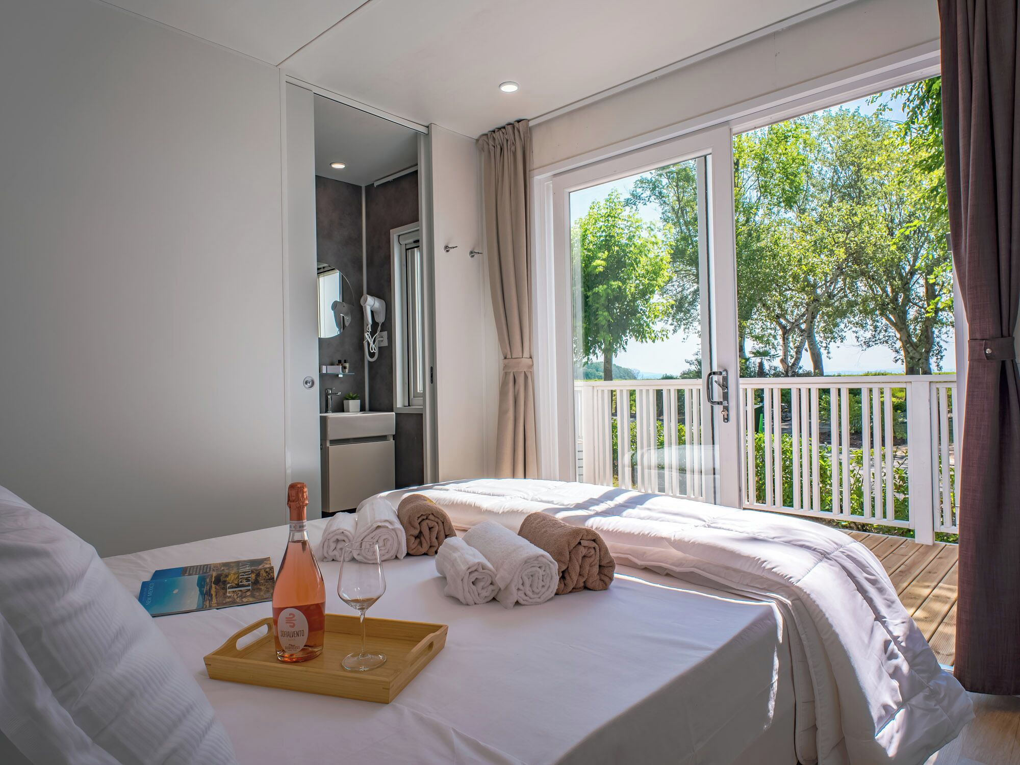 A hotel room with a balcony, a bed, a bathroom, and a tray with a bottle and glass.