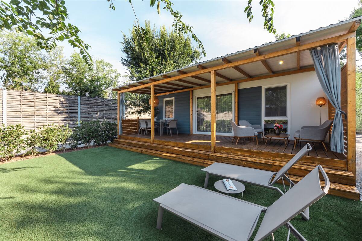 A covered outdoor patio with a wooden deck, a seating area with two lounge chairs and a table, and a view of the surrounding garden and trees.