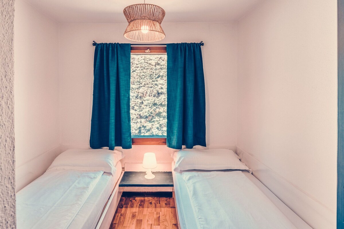 A small room with two beds, a window with curtains, a wooden floor, and a hanging light fixture.