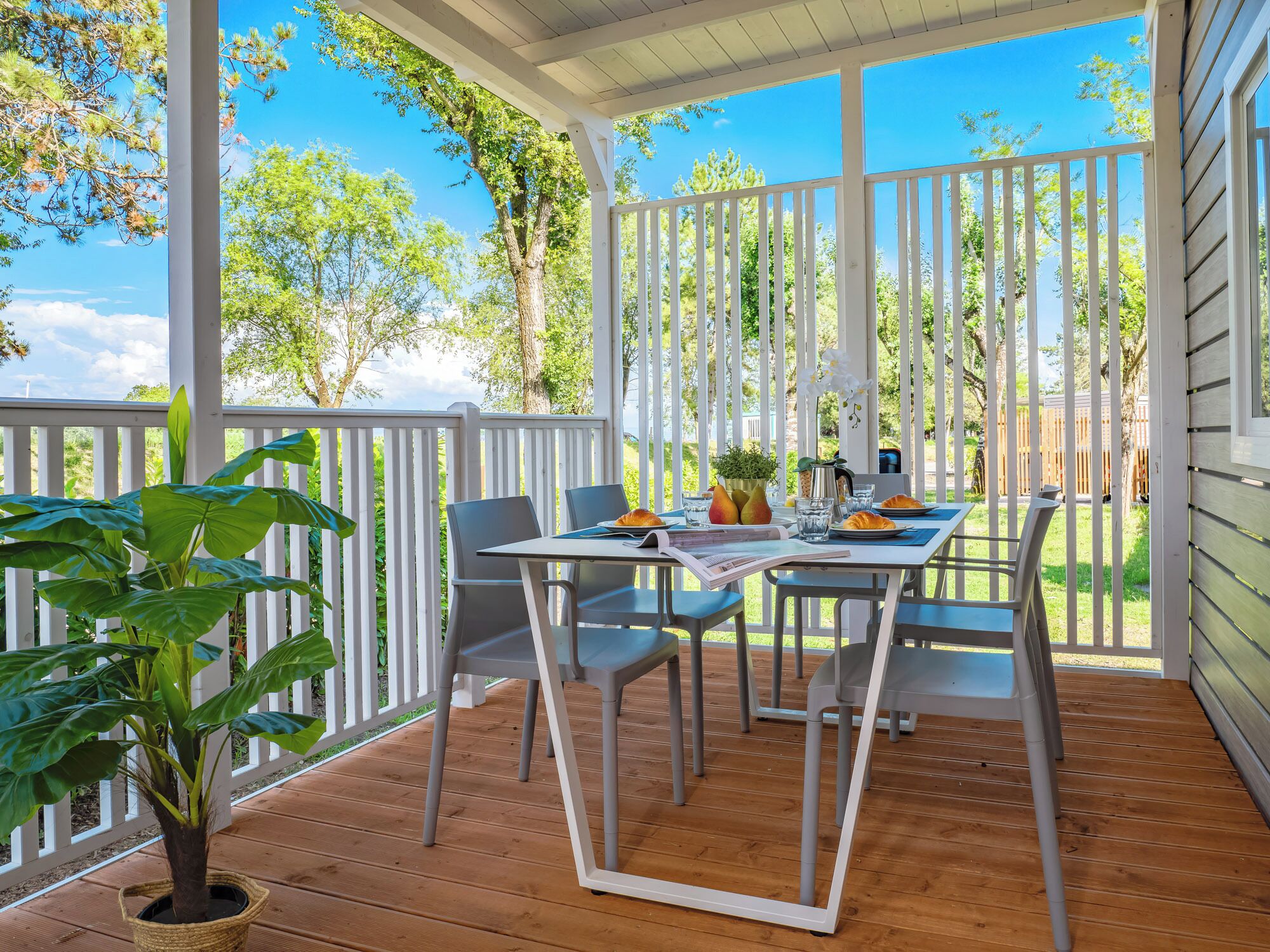 A covered outdoor patio with a table and chairs, surrounded by white railings and greenery.