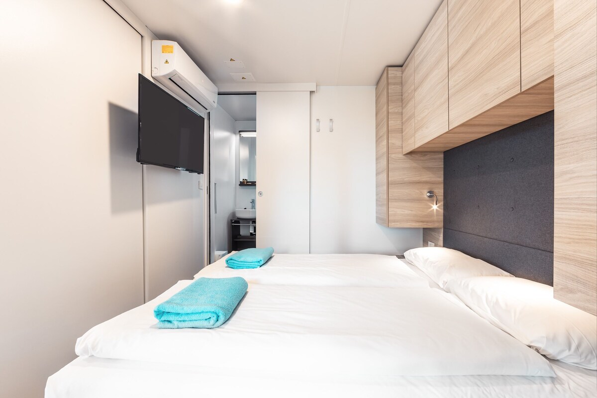 Une chambre d’hôtel moderne avec deux lits, un téléviseur à écran plat fixé au mur et une armoire intégrée dotée d’une tête de lit grise.