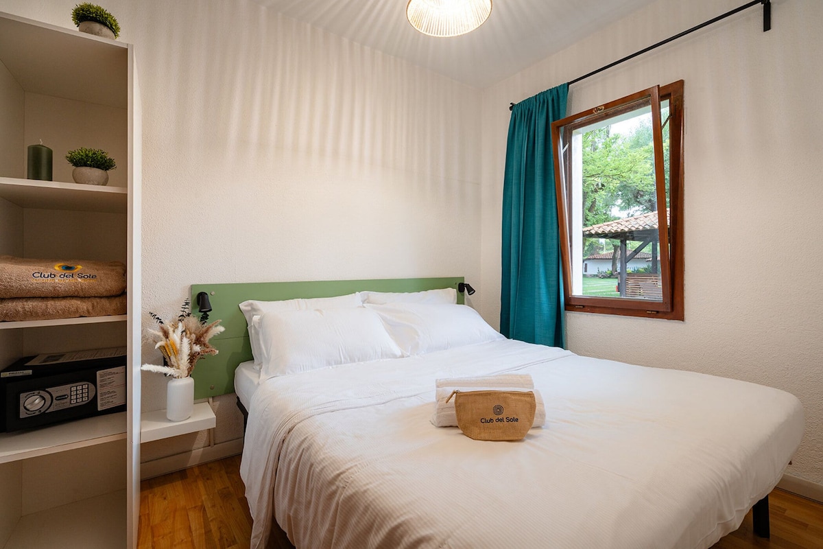 Un lit bien fait, avec une couette blanche et des oreillers, une table de chevet en bois avec une lampe, une étagère avec des serviettes et une petite plante, et une fenêtre donnant sur un paysage verdoyant.
