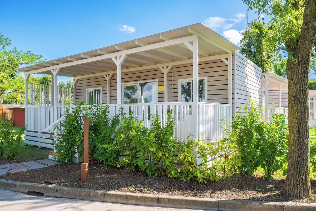 Einbungalow mit Veranda, umgeben von viel Grün und blauem Himmel.