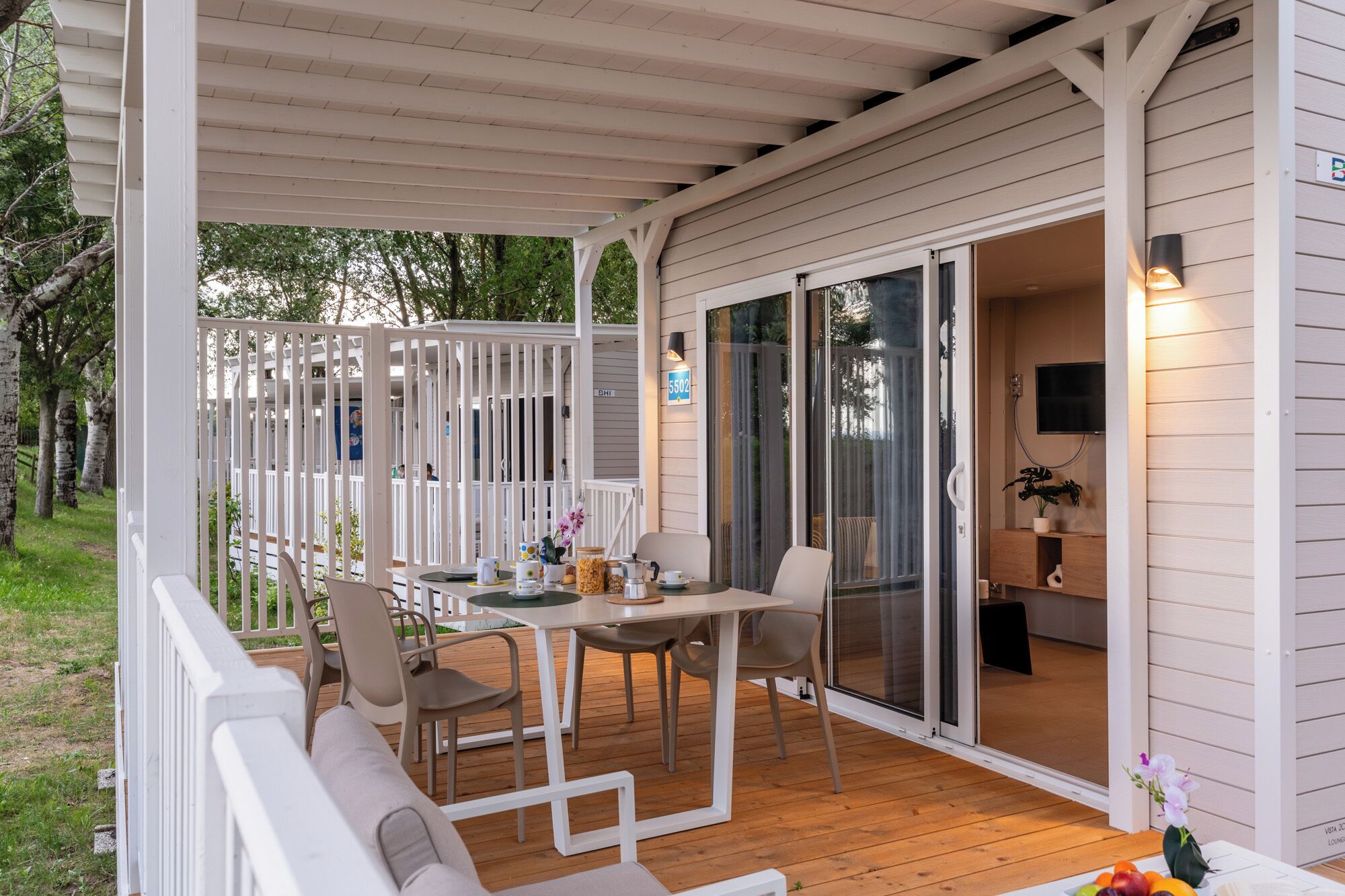 A covered outdoor seating area with a table and chairs, a sofa, and a sliding glass door leading to an interior space with a TV and a plant.