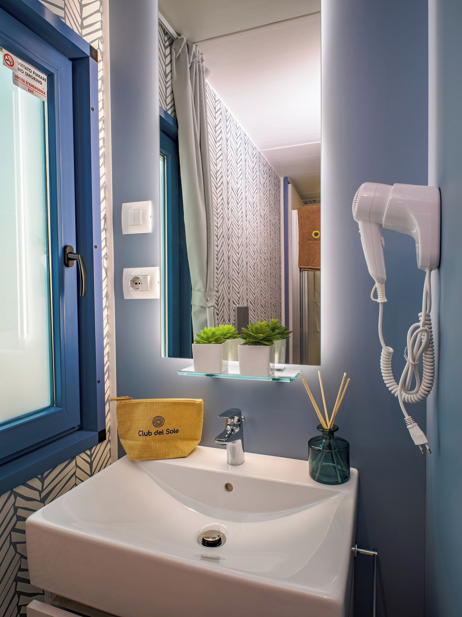 Ein Badezimmer mit einem weißen Waschbecken, einem an der Wand montierten Föhn, einem Handtuchhalter und einem Fenster mit blauem Rahmen.