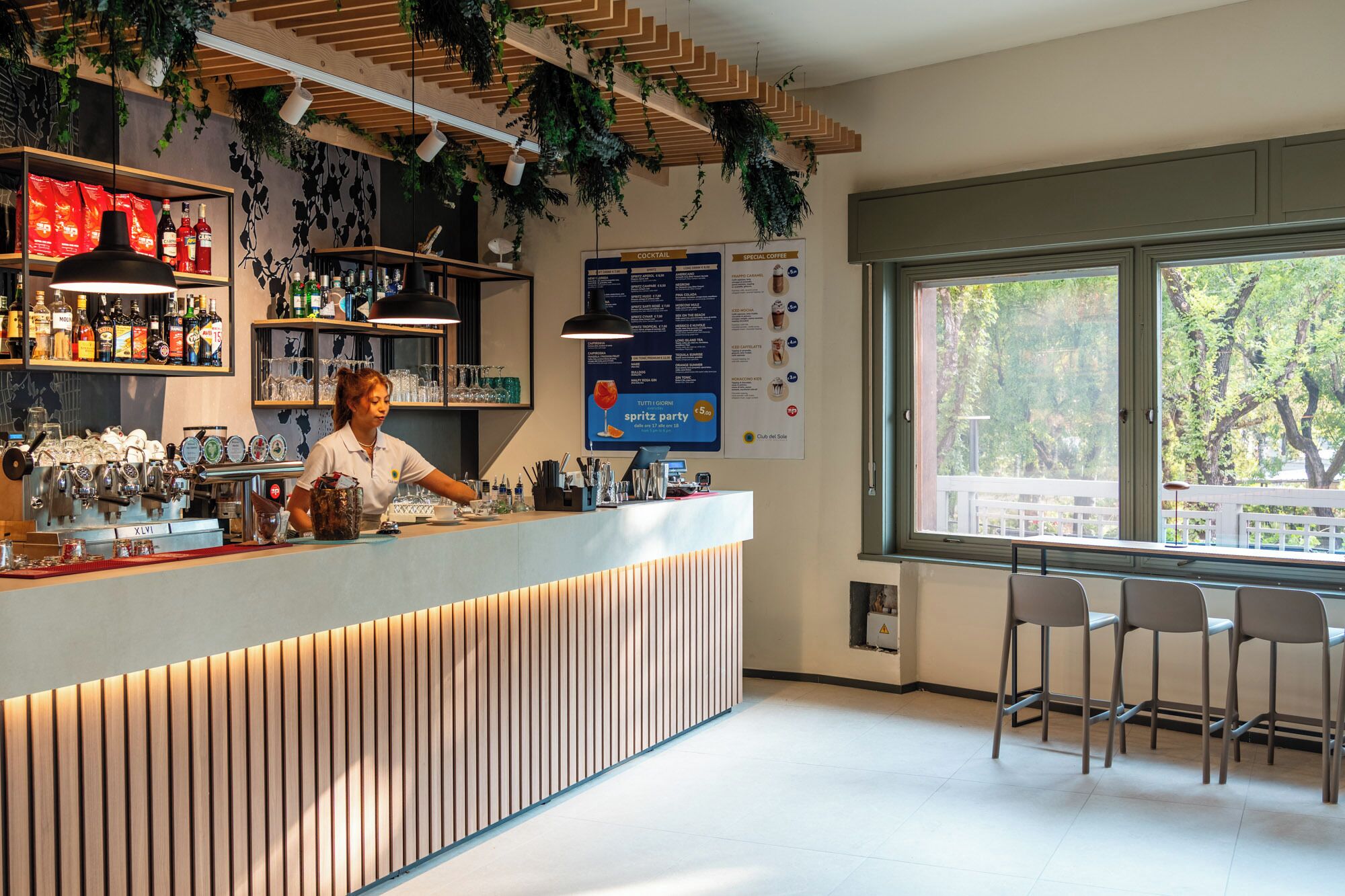 Zona de bar amb un barman, estantes plens de garrafes, un tauler amb el menú i una vista per la finestra.