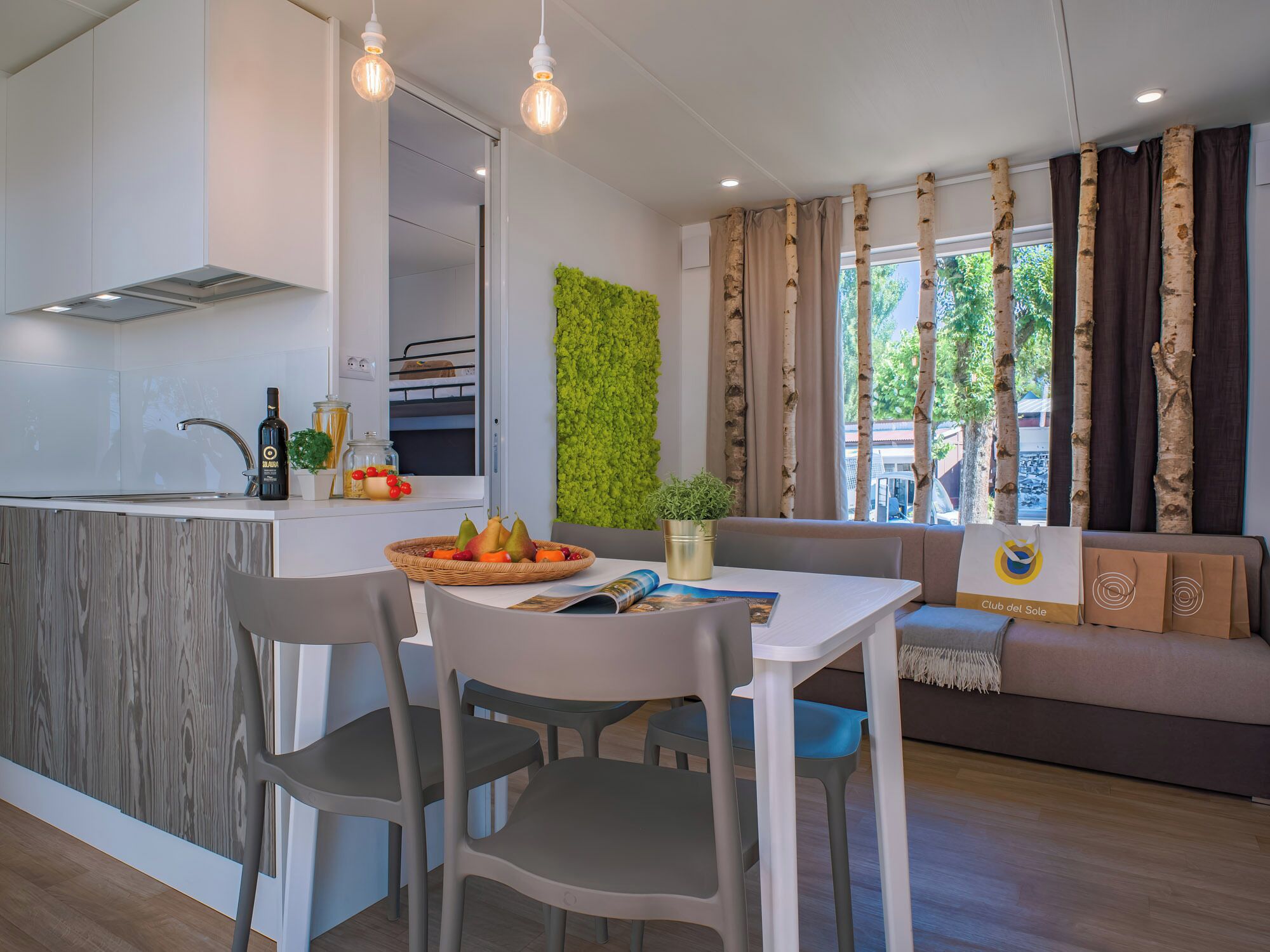 A modern kitchen with a built-in island, a dining area with a table and chairs, a sofa, and a view of trees through large windows.