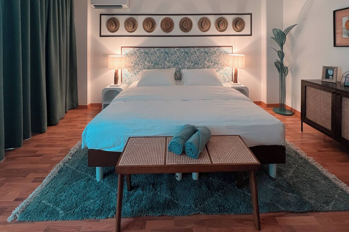 Ein ordentlich eingerichtetes Hotelzimmer mit einem großen Bett, zwei Nachttischen, einer Stehlampe, einer wandmontierten Klimaanlage und einem gerahmten Kunstwerk über dem Bett.