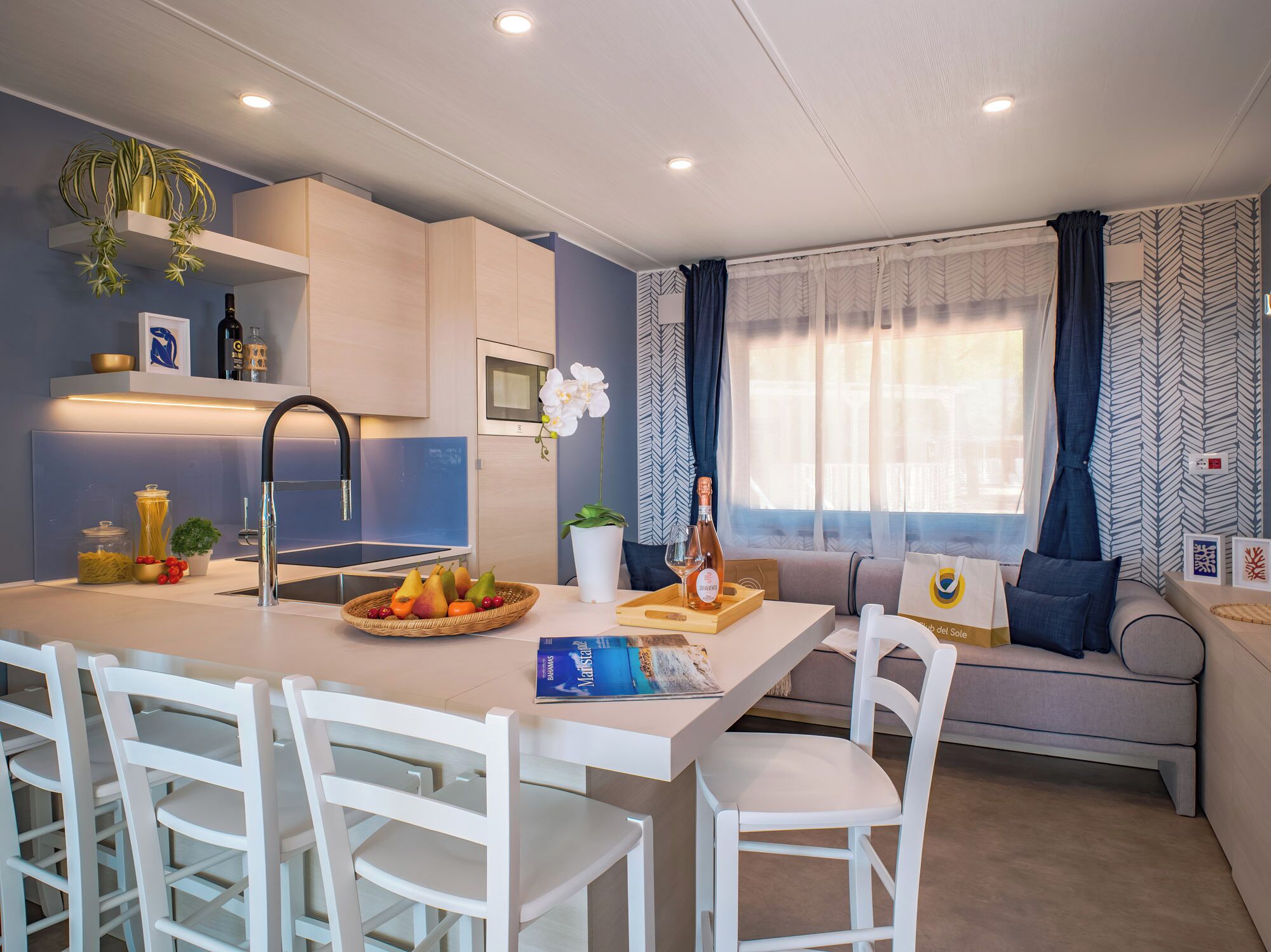 A modern kitchen with a central island, white chairs, a dining table, and a sofa with cushions.