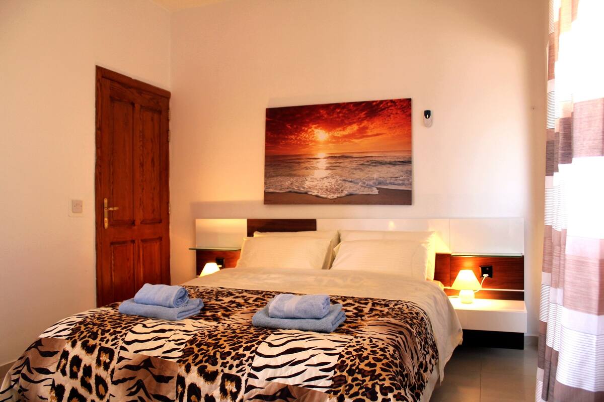 A bedroom with a bed, a painting of a sunset over the ocean, and a wooden door.