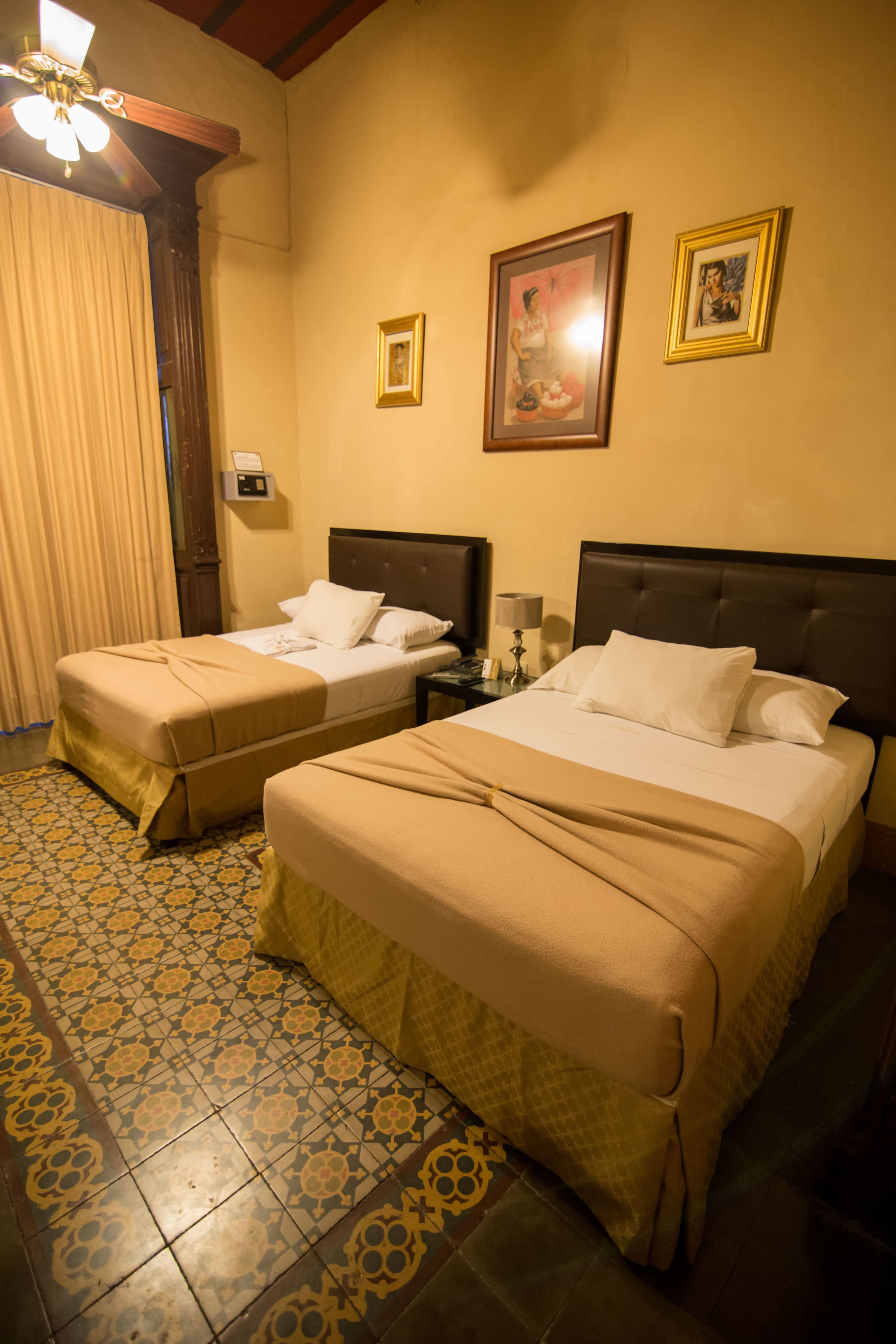 Habitación de hotel con dos camas, ventilador de techo, mesita de noche con lámpara y un reloj en la pared.