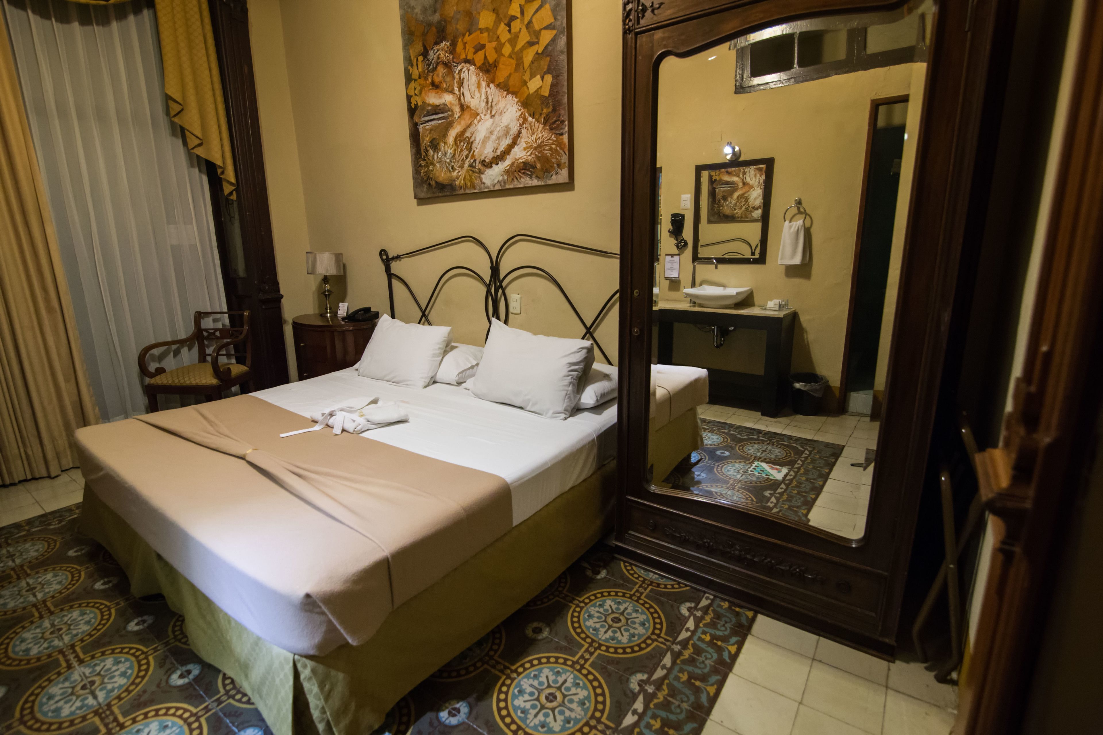A hotel room with a bed, a bedside table with a lamp, a bathroom mirror, and a patterned floor.