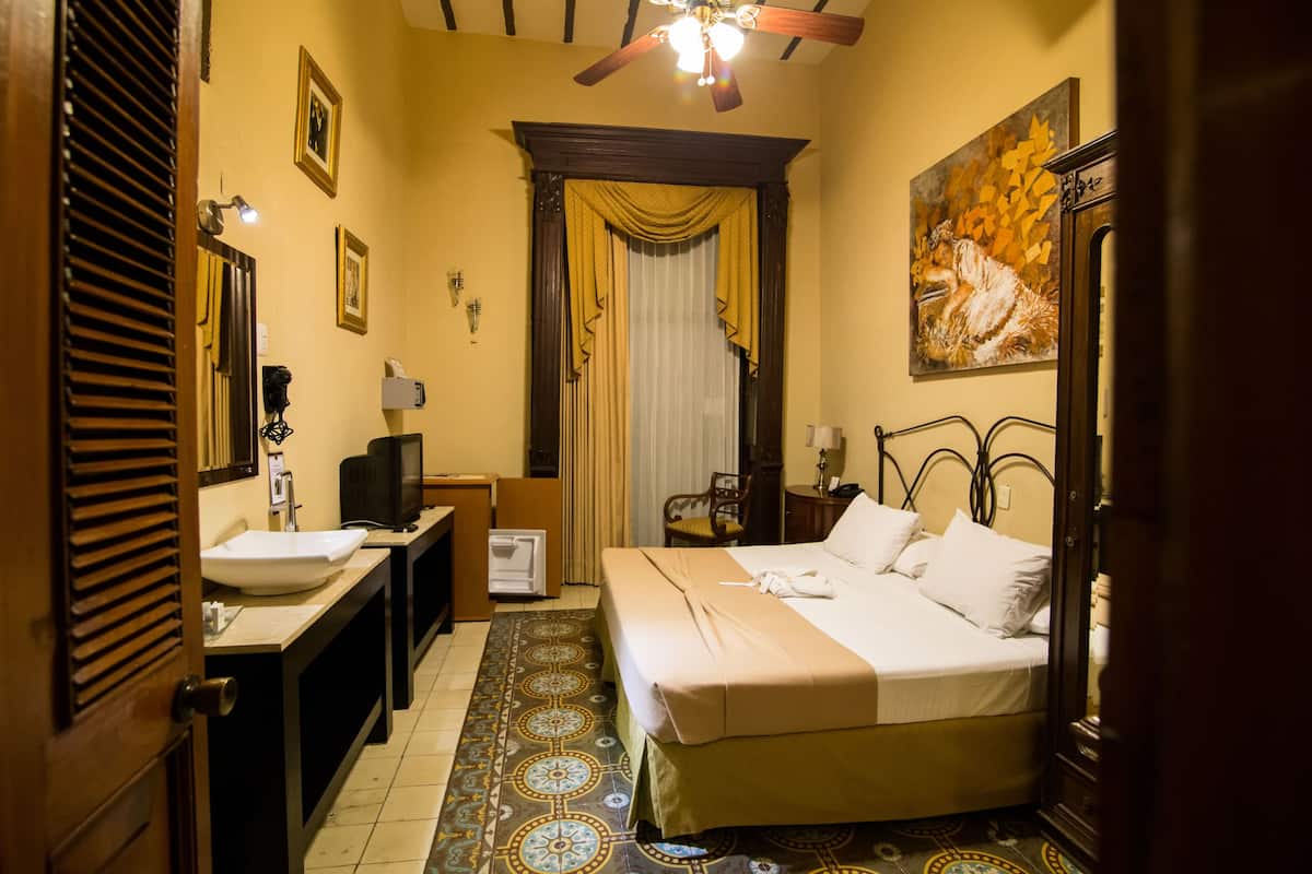Habitación de hotel con cama, lavamanos, espejo, televisor y un cuadro en la pared.