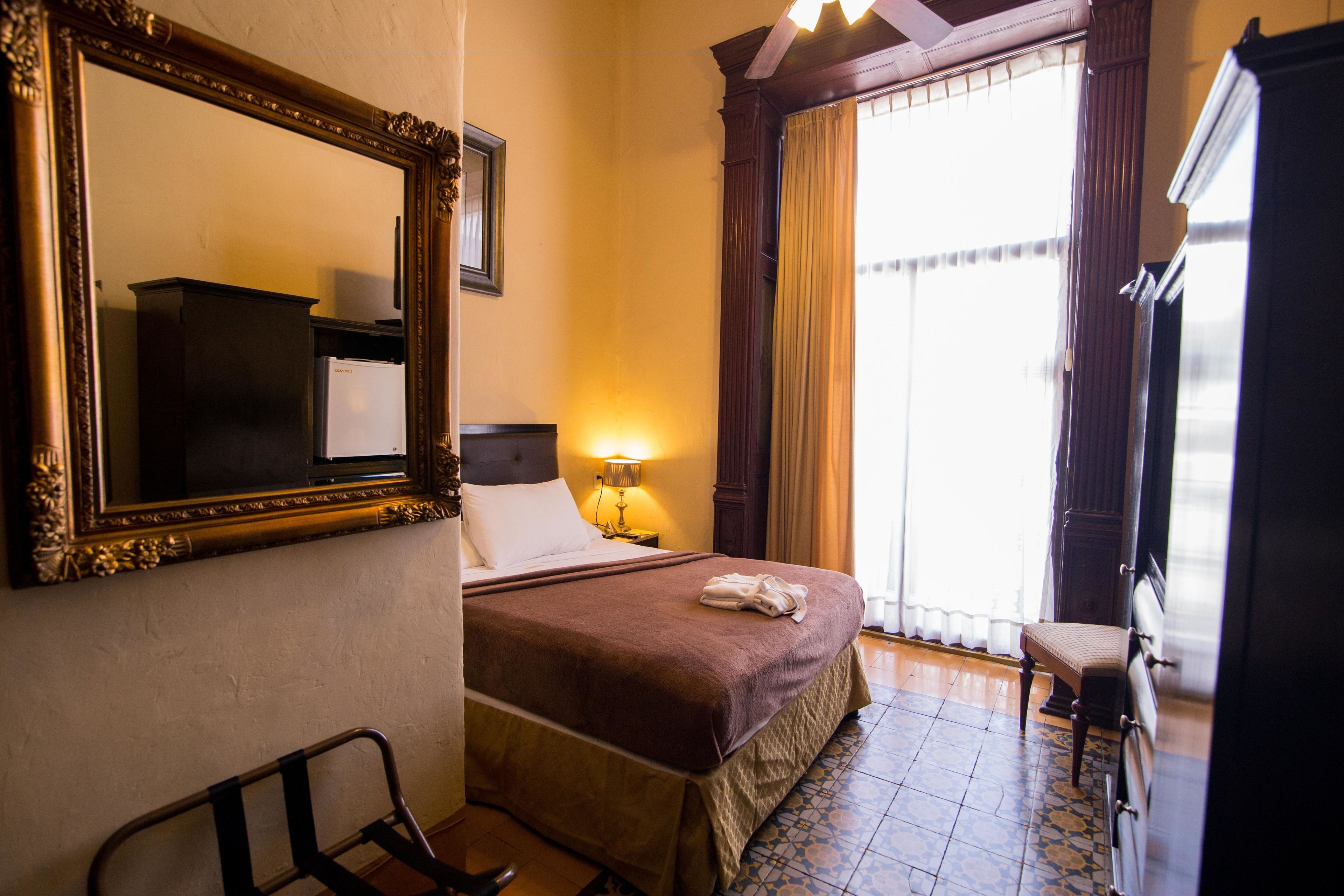 A hotel room with a large bed, a bedside table with a lamp, a chair, a wardrobe, and a large mirror on the wall.