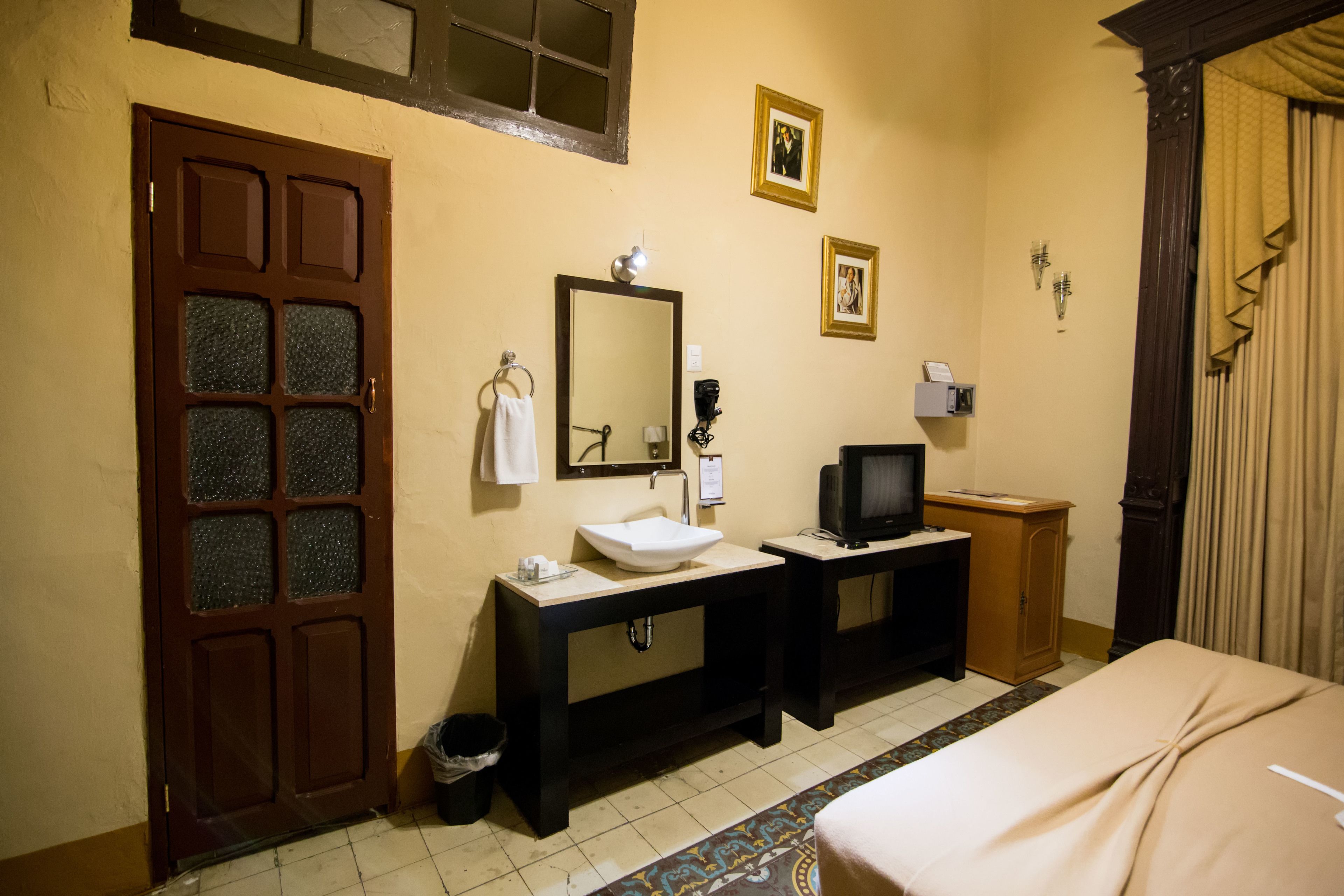 A hotel room with a wooden door, a large mirror, a sink, a television, and a bed.