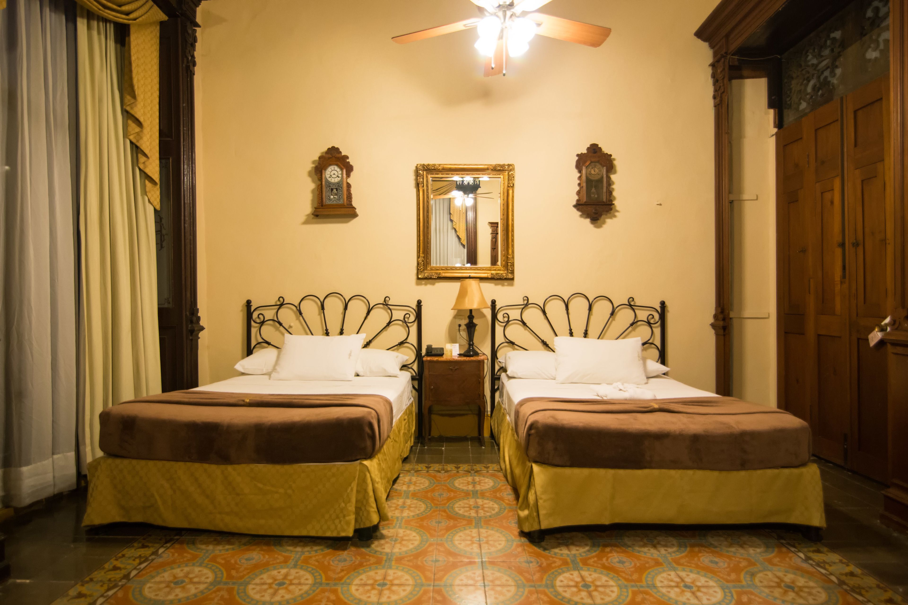 A room with two single beds, a ceiling fan, a mirror, and a clock on the wall.