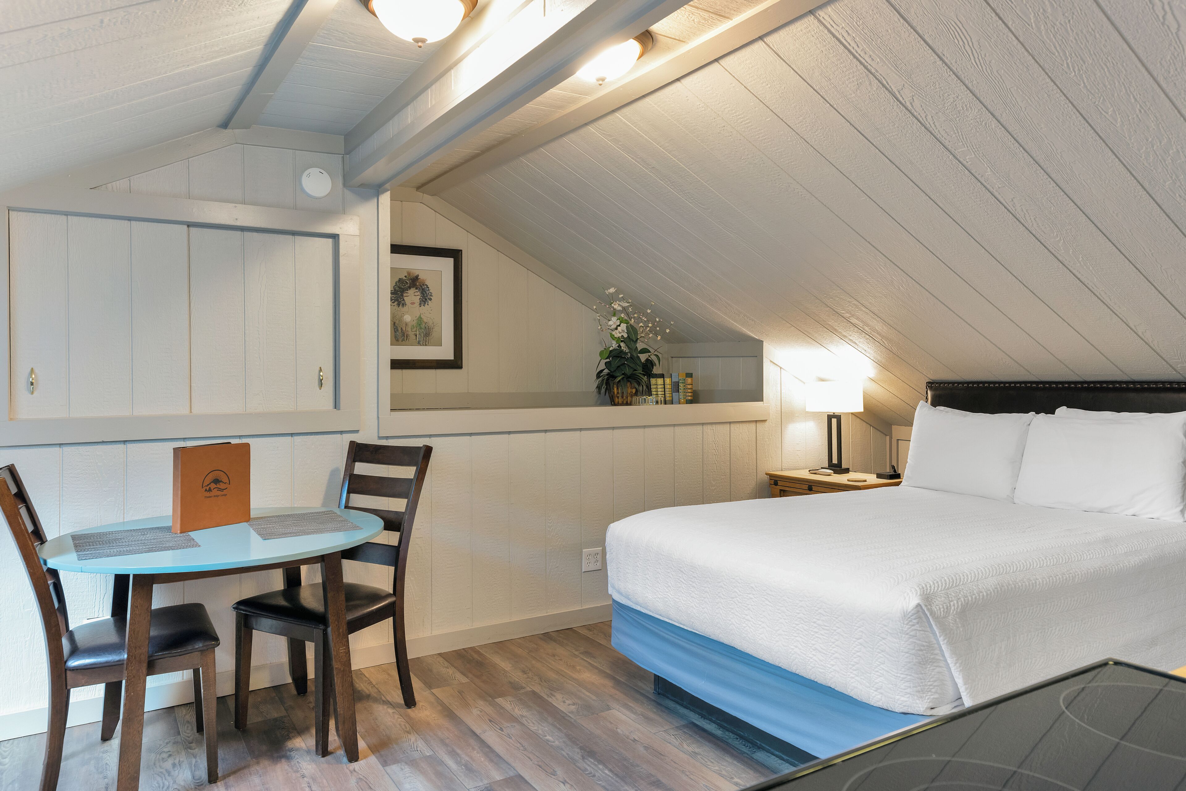 A bedroom with a bed, a desk with chairs, a lamp, and a small table with a plant.