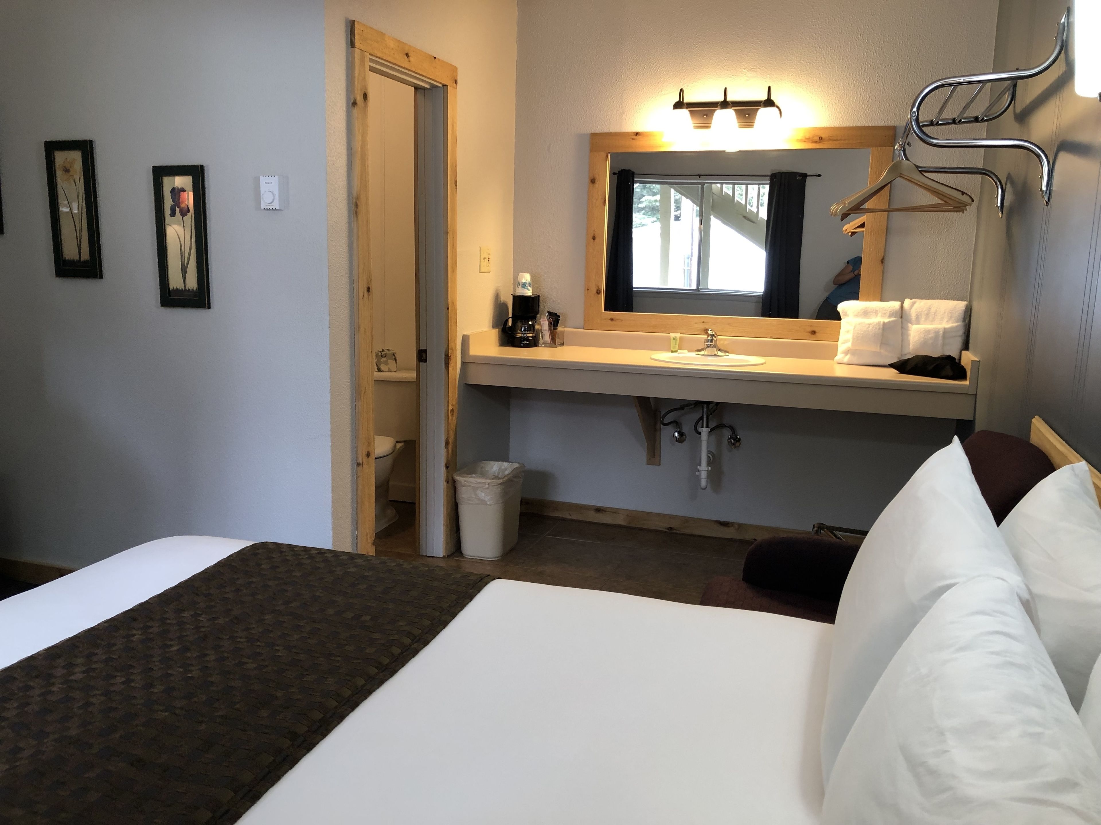 Camera d'albergo con un letto, un bagno con lavandino e specchio, e un corridoio che porta a un'altra stanza.