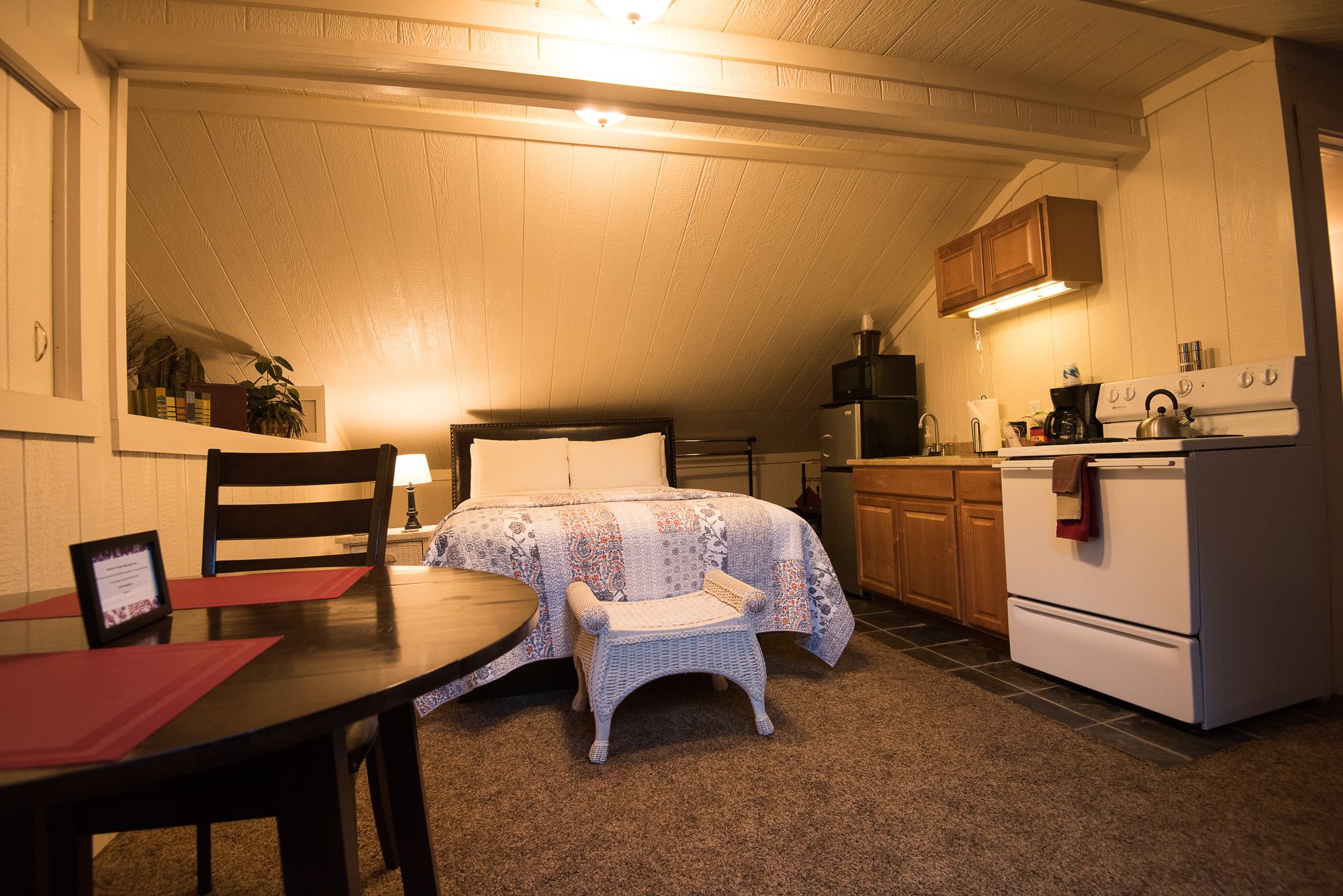 A compact, well-lit room with a bed, a small dining table, a kitchen area with a stove and refrigerator, and a chair.