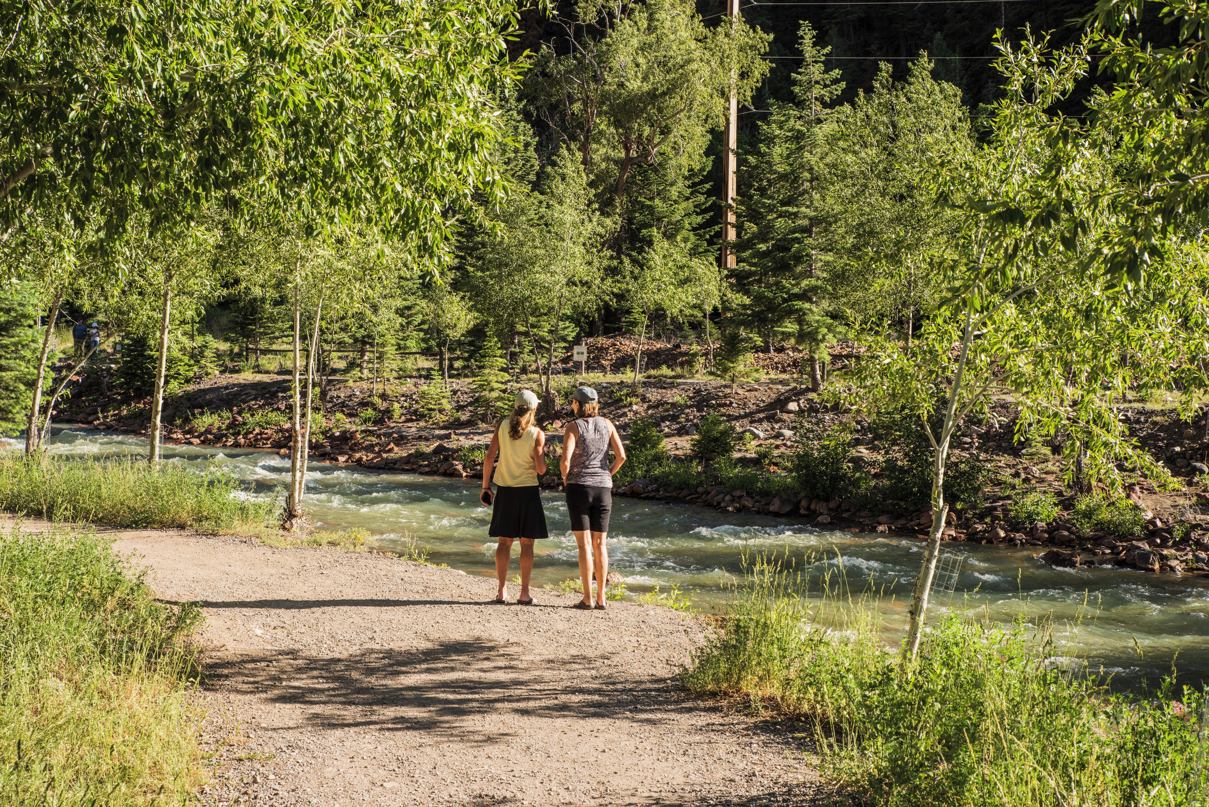 Two people standing on a dirt path by a river with trees and a fence in the background.