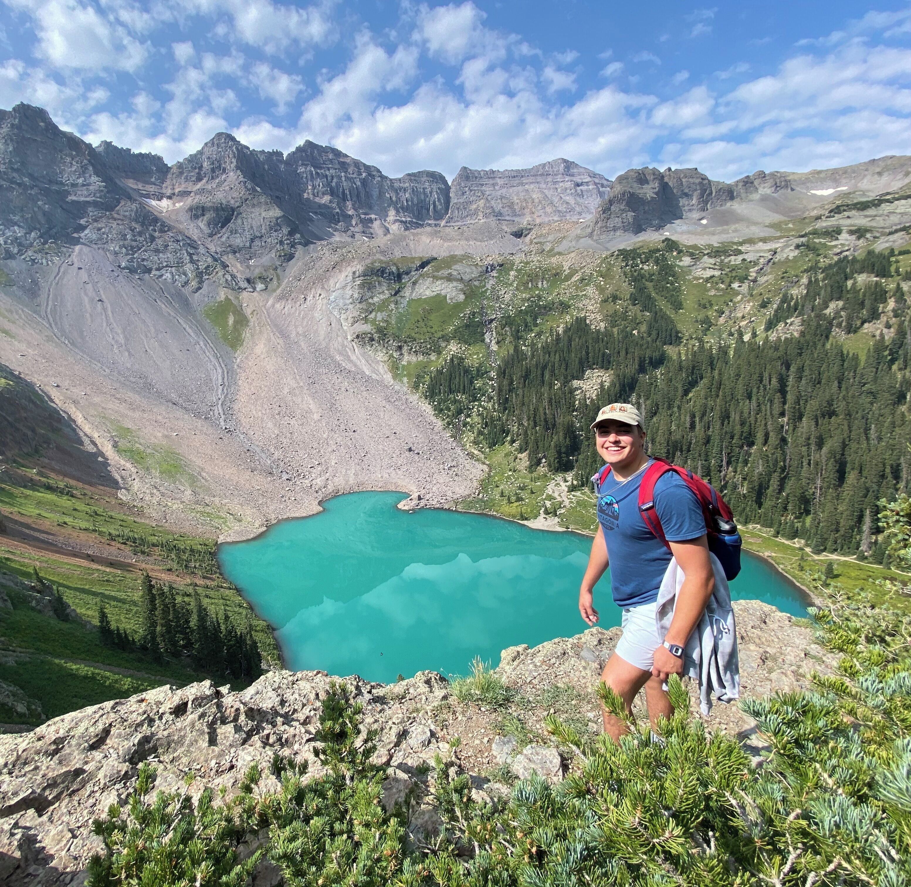 A hiker on a mountain trail with a lake and forested mountains in the background.