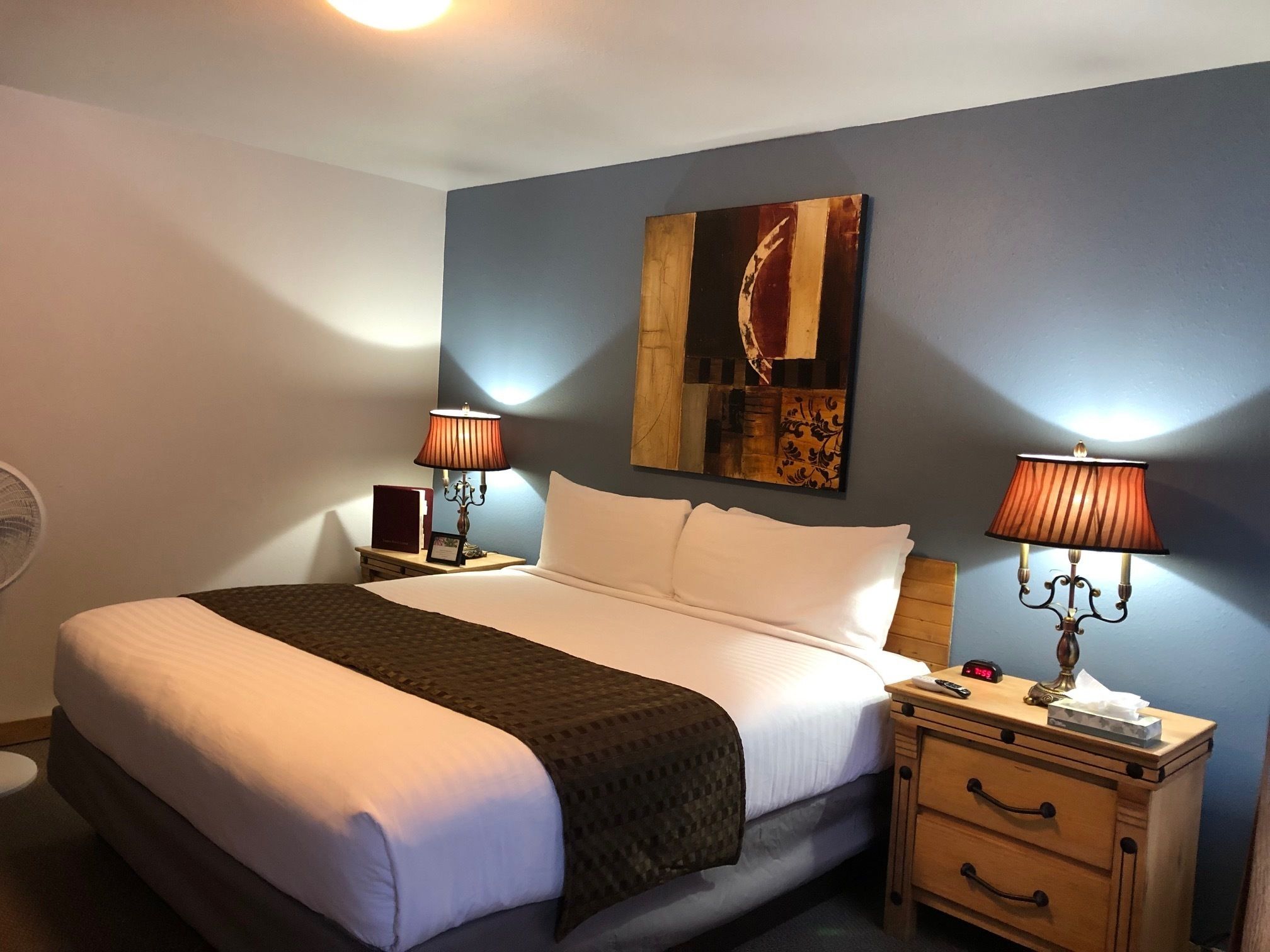 A neatly made bed with a white comforter and a dark throw blanket, flanked by two bedside tables with lamps, a wall-mounted artwork, and a wooden nightstand with a clock and a tissue box.