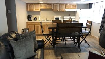 A compact living space with a kitchenette, wooden cabinets, a dining table with chairs, a leather sofa, and a small wooden coffee table.