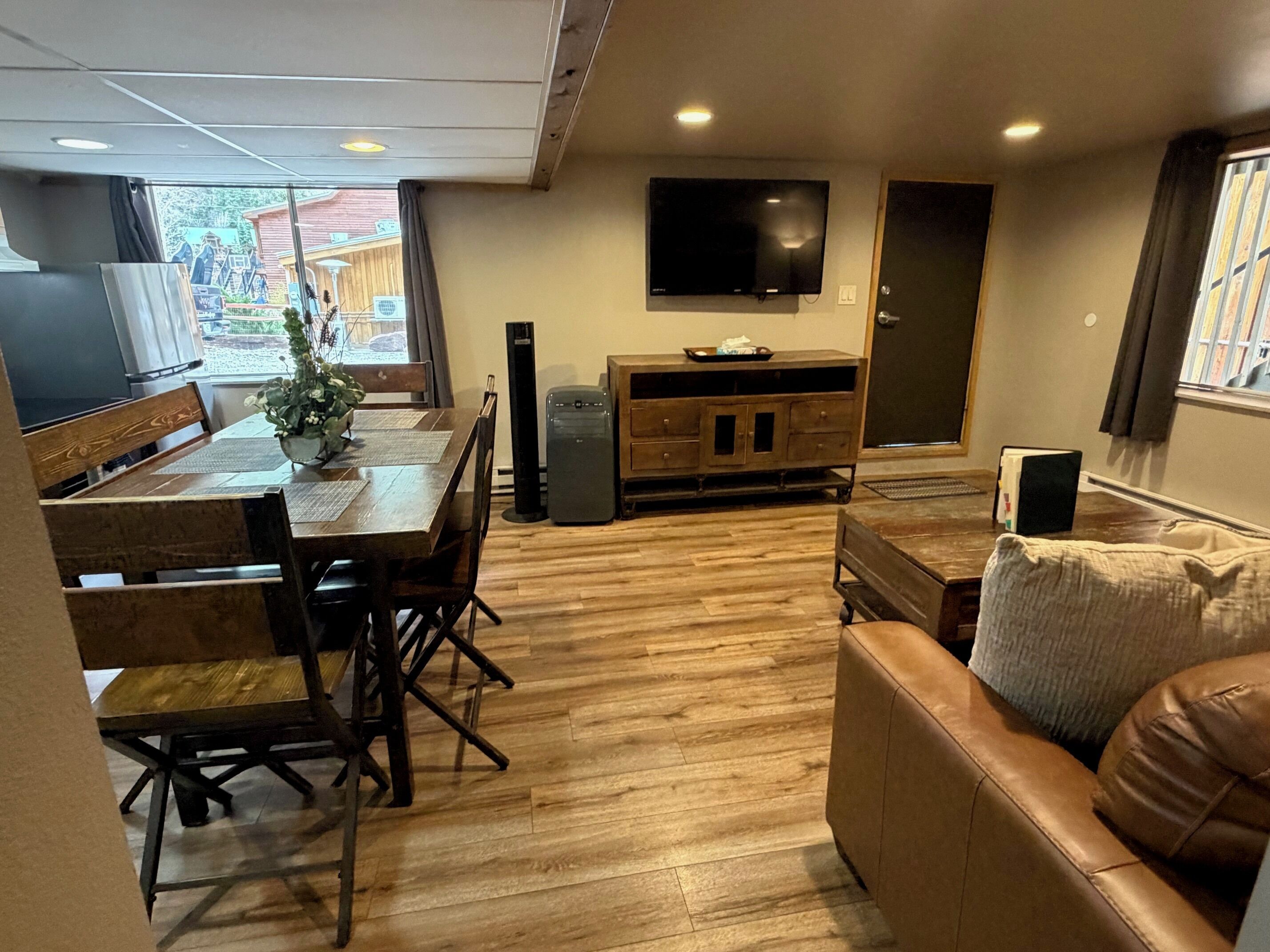 A living room with a wooden floor, a sofa, a dining table with chairs, a TV mounted on the wall, and a window with curtains.