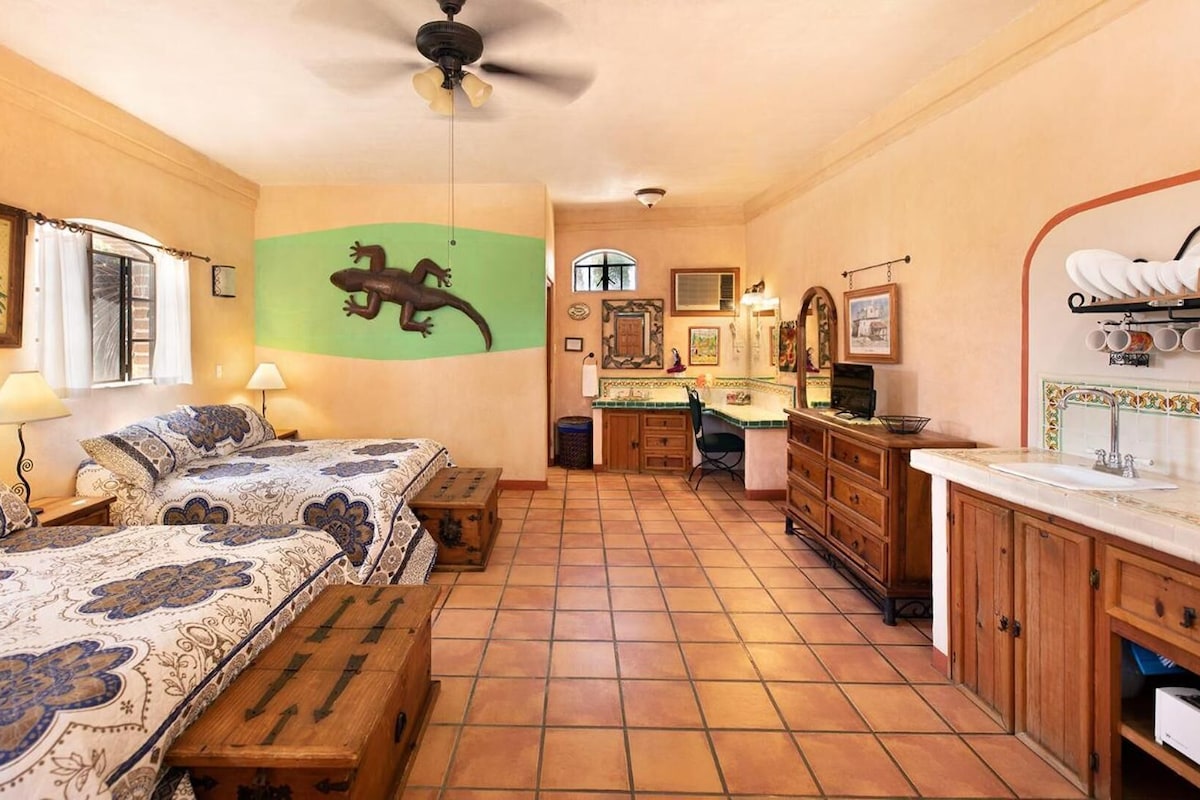 A cozy room with a bed, a bench, a ceiling fan, a kitchen area with a sink, and a green wall decoration.