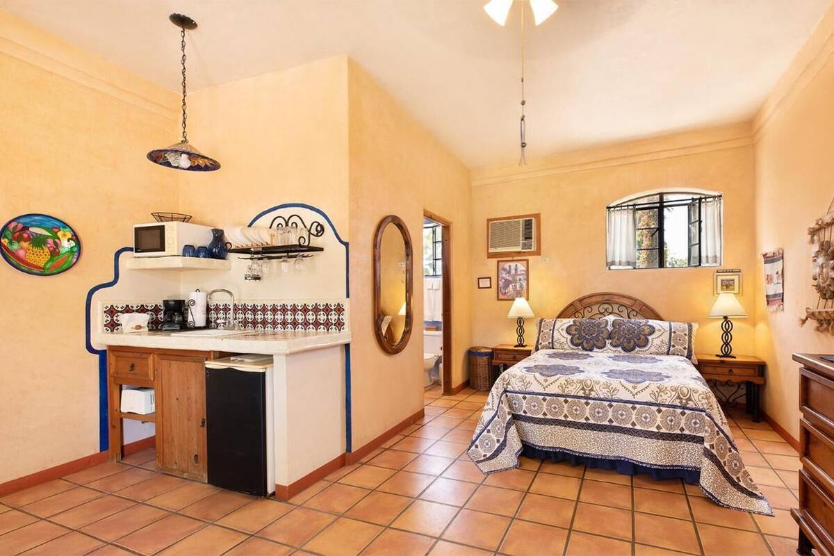 A compact bedroom with a bed, bedside table, lamp, and a TV. There is a kitchenette with a microwave, kettle, and small refrigerator. The room has a tiled floor and a window with a view.